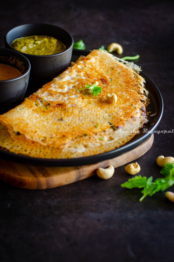 crispy Instant rava dosa served with chutney by the side on a black plate. Coriander leaves and cashews sprinkled on the top and by the side.