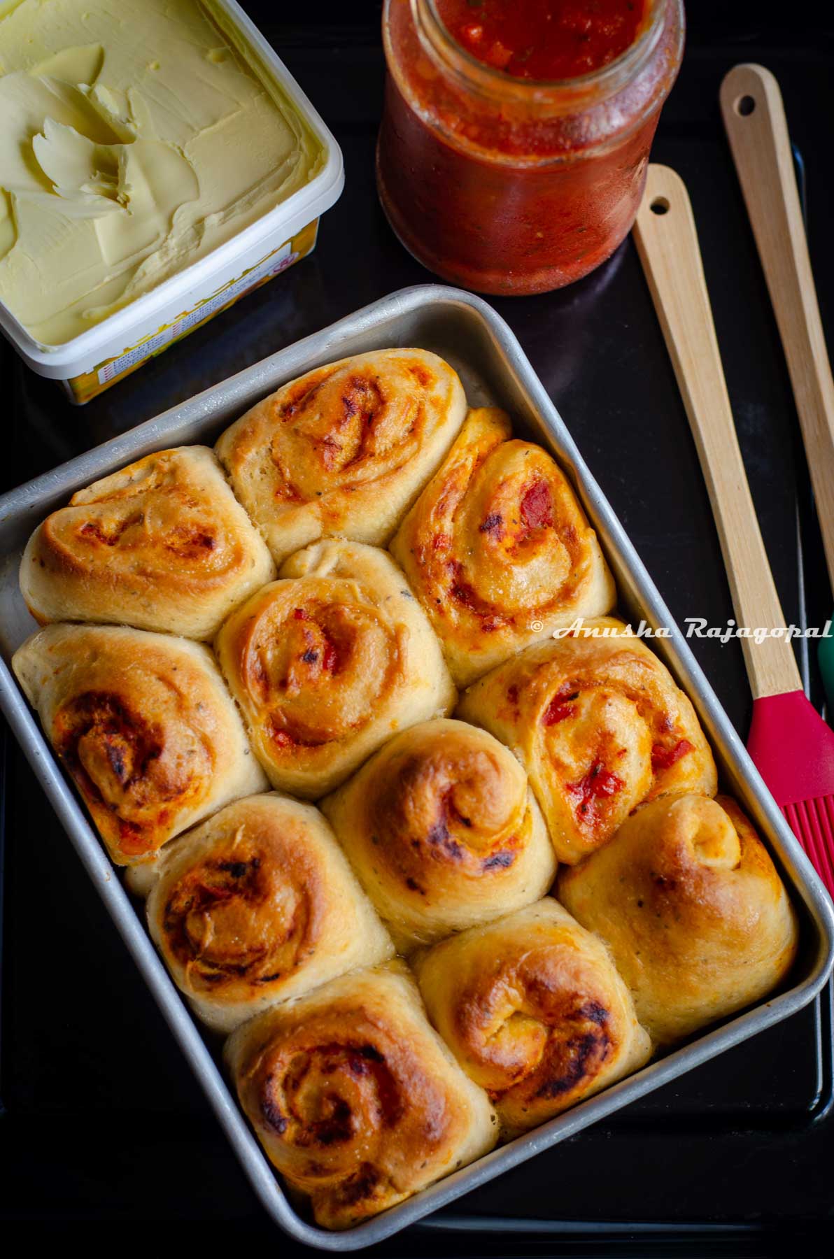 freshly baked pull apart rolls filled with store bought pasta sauce in a baking tray. Spatulas, a jar of sauce and some vegan butter by the side.