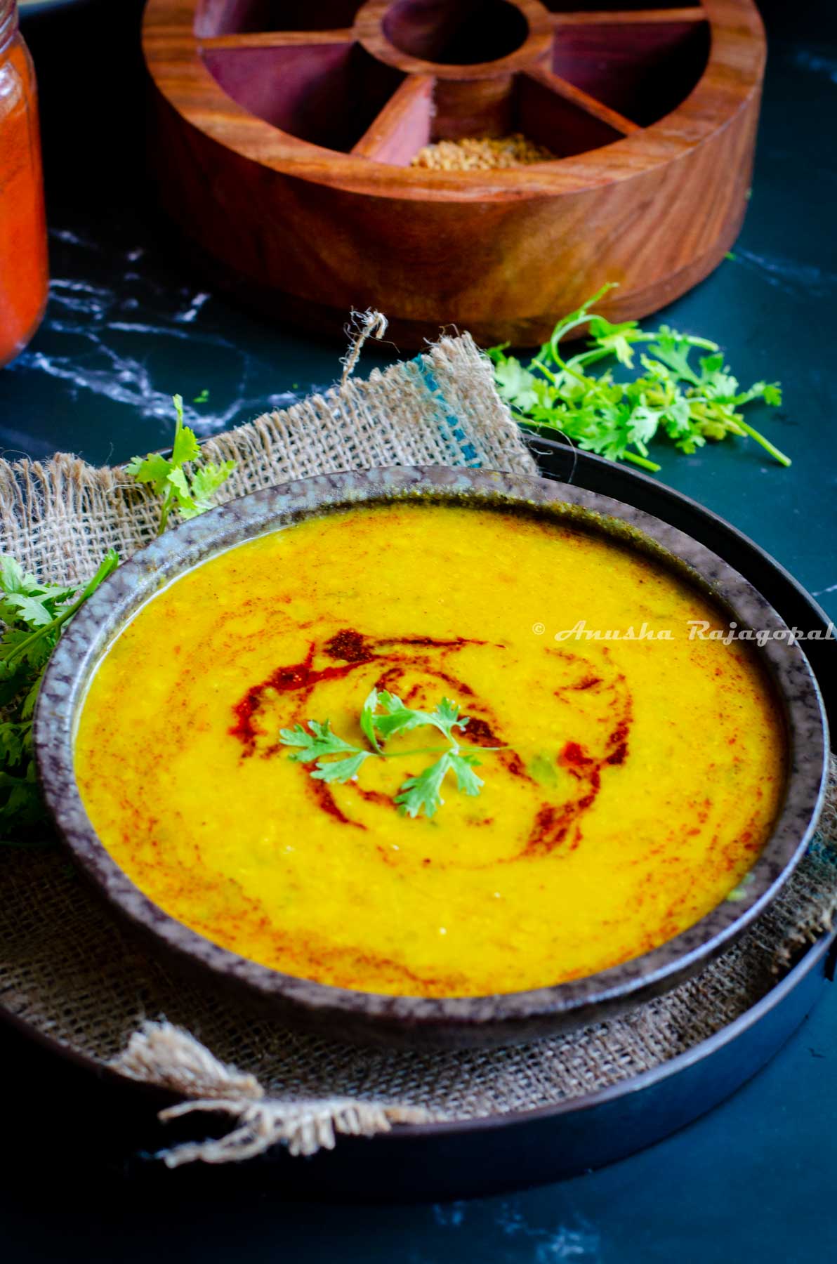 dal tadka topped with a tempering and garnished with finely chopped cilantro leaves. Dal served in a black bowl placed on a burlap mat. Spice box and spices at the background.