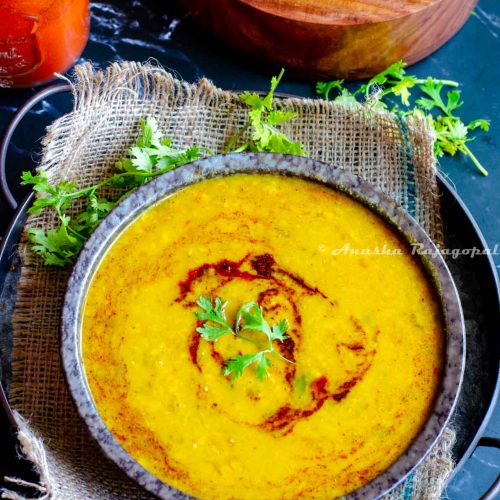 dal tadka topped with a tempering and garnished with finely chopped cilantro leaves. Dal served in a black bowl placed on a burlap mat. Spice box and spices at the background.