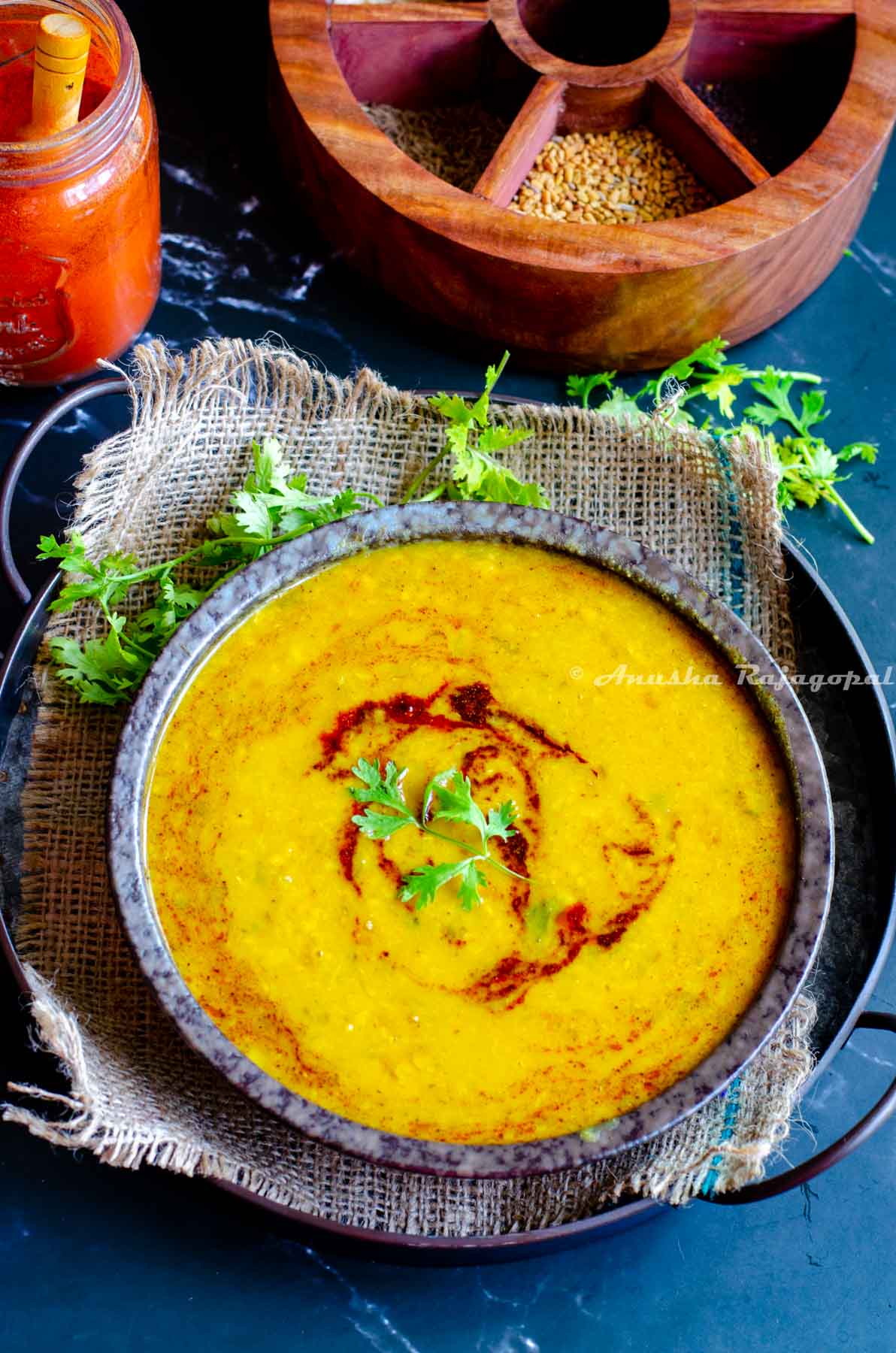 dal tadka topped with a tempering and garnished with finely chopped cilantro leaves. Dal served in a black bowl placed on a burlap mat. Spice box and spices at the background.