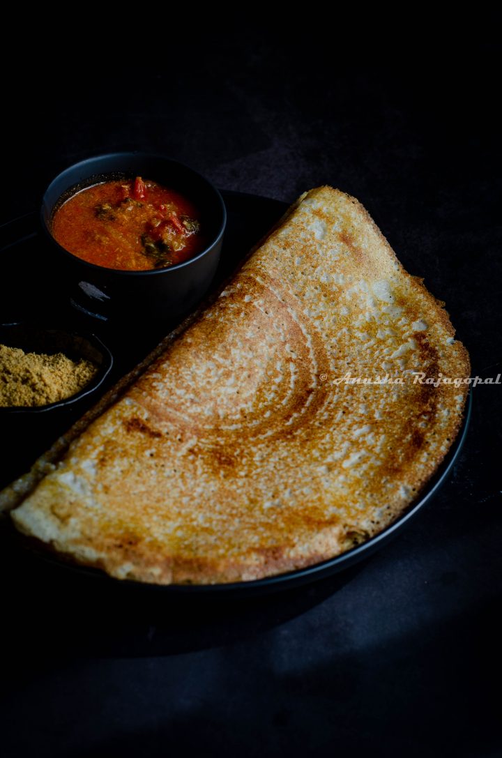 crispy barley dosas served on a black plate with a spicy chutney and Idli podi as accompaniments