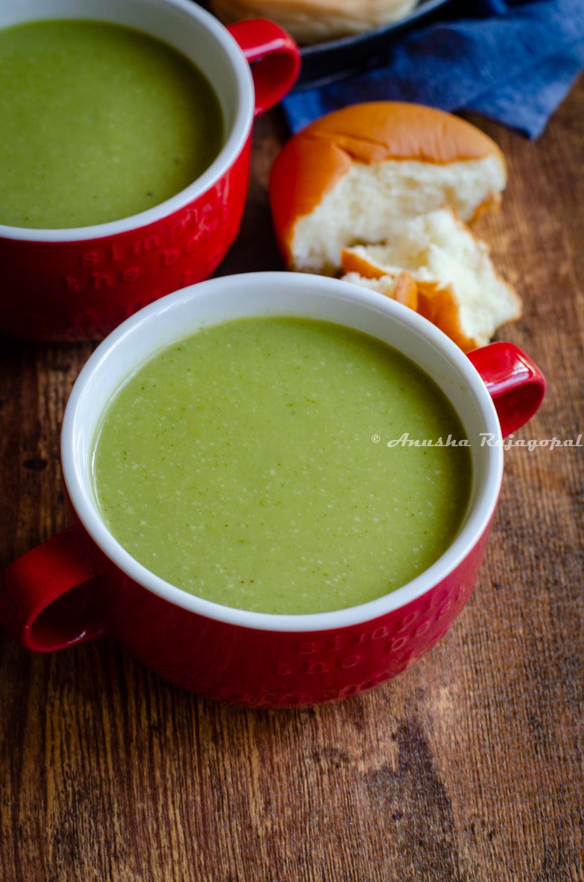 vegan broccoli almond soup served in a red soup bowl with a half eaten roll at the back.