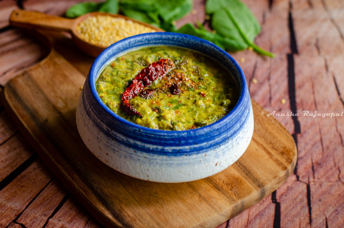 keerai kootu served in a white and blue bowl placed over a wooden chopping board. Moong lentils and spinach leaves by the side.