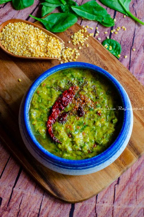 keerai kootu served in a white and blue bowl placed over a wooden chopping board. Moong lentils and spinach leaves by the side.