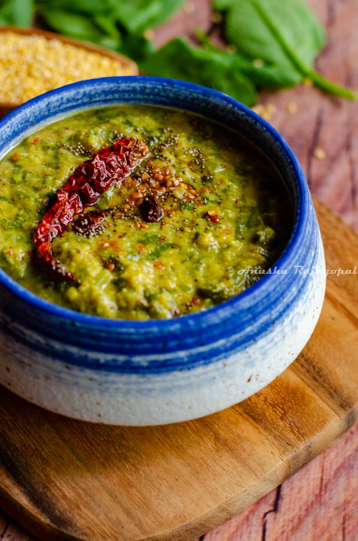 keerai kootu served in a white and blue bowl placed over a wooden chopping board. Moong lentils and spinach leaves by the side.