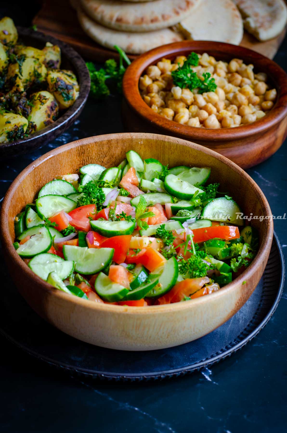 Persian Shirazi salad served in a wooden salad bowl placed over a metal serving platter. Hummus Balila, Batata Harra at the background