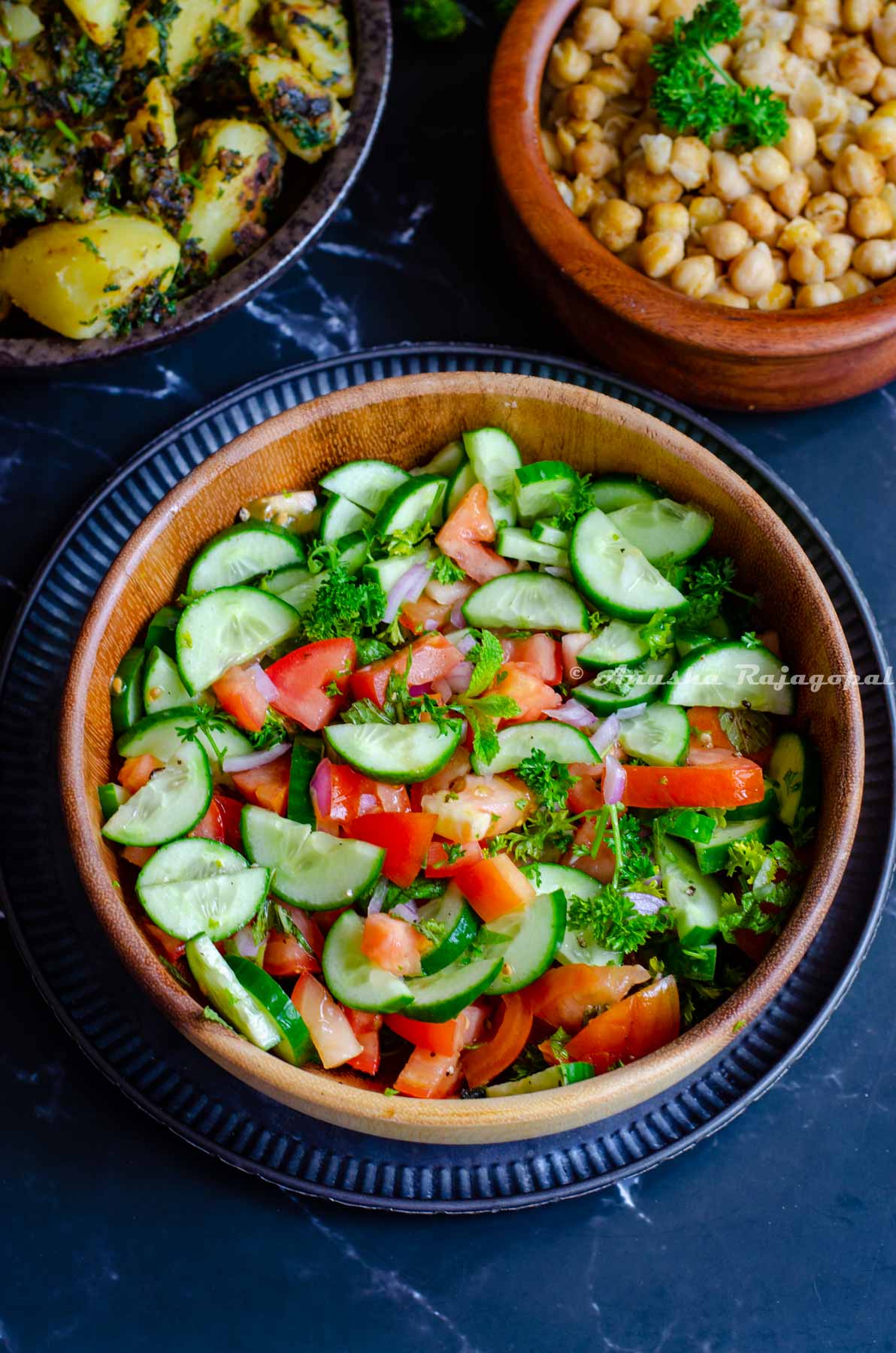 Persian Shirazi salad served in a wooden salad bowl placed over a metal serving platter. Hummus Balila, Batata Harra at the background