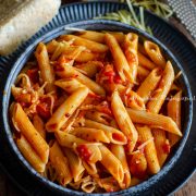 Penne arrabiata served in a greyish black shallow plate with herbs and slices of bread by the side. A cheese grater is also featured alongside.