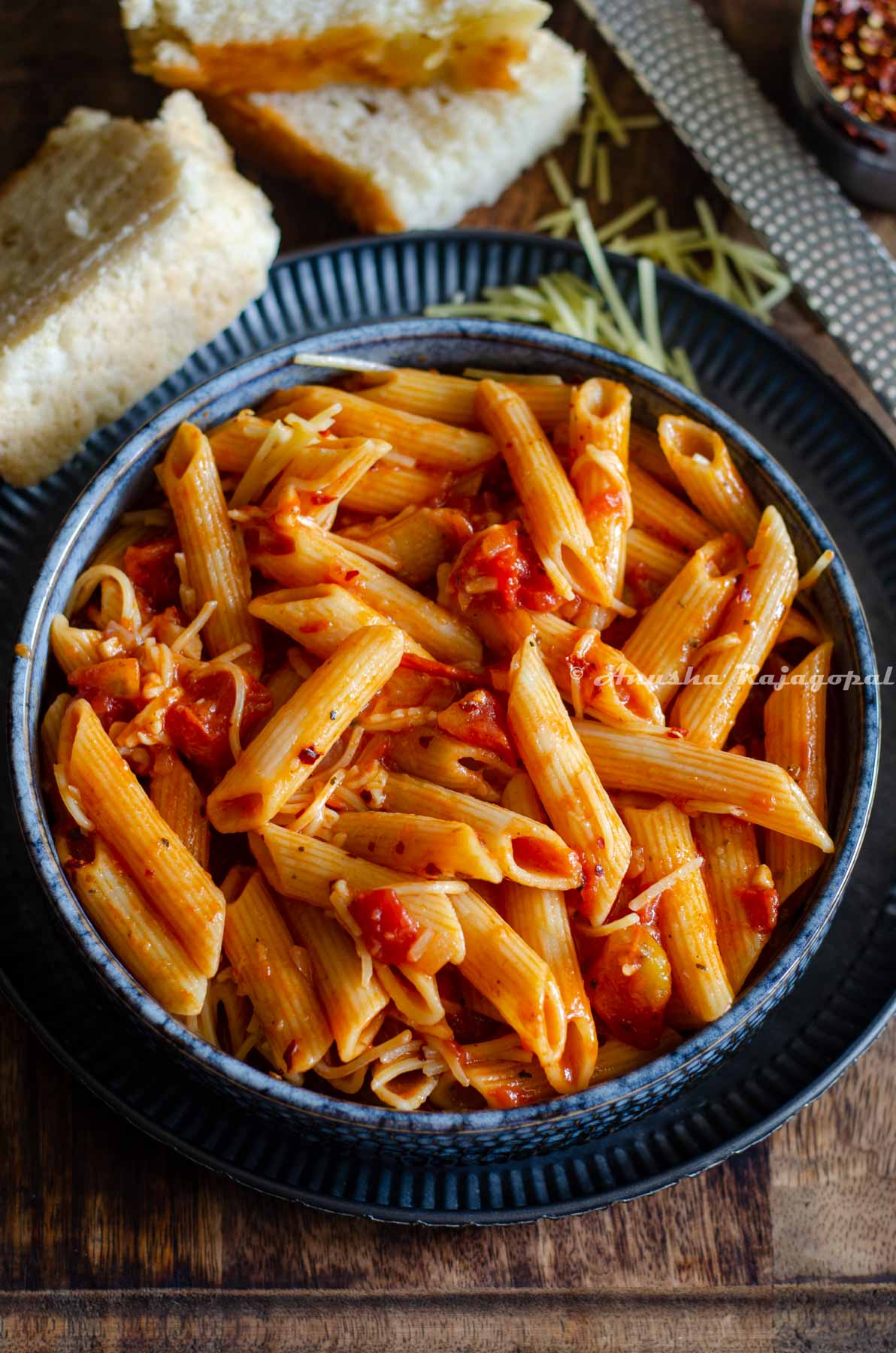 Penne arrabiata served in a greyish black shallow plate with herbs and slices of bread by the side. A cheese grater is also featured alongside.