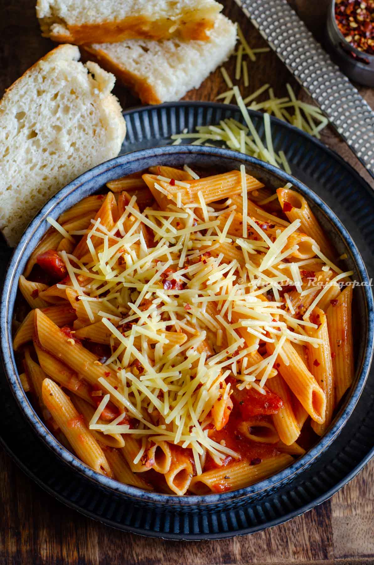 Arrabiata pasta topped with grated parmesan and chili flakes, served in a shallow black bowl. Home baked bread, a cheese grated and a small jar of chili flakes at the back ground 