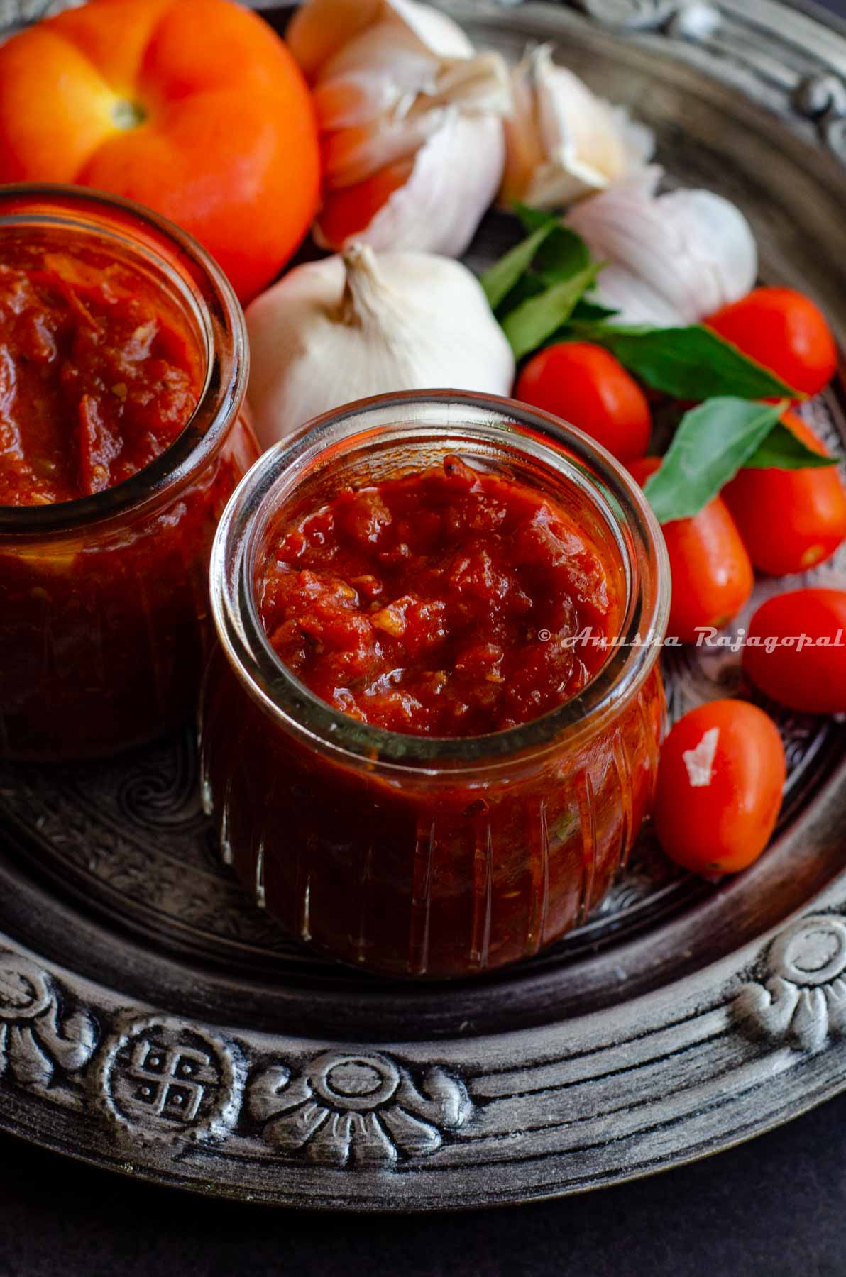 South Indian thakkali thokku- a tangy tomato relish in a glass jar with cherry tomatoes, garlic and curry leaves all presented in a rustic platter