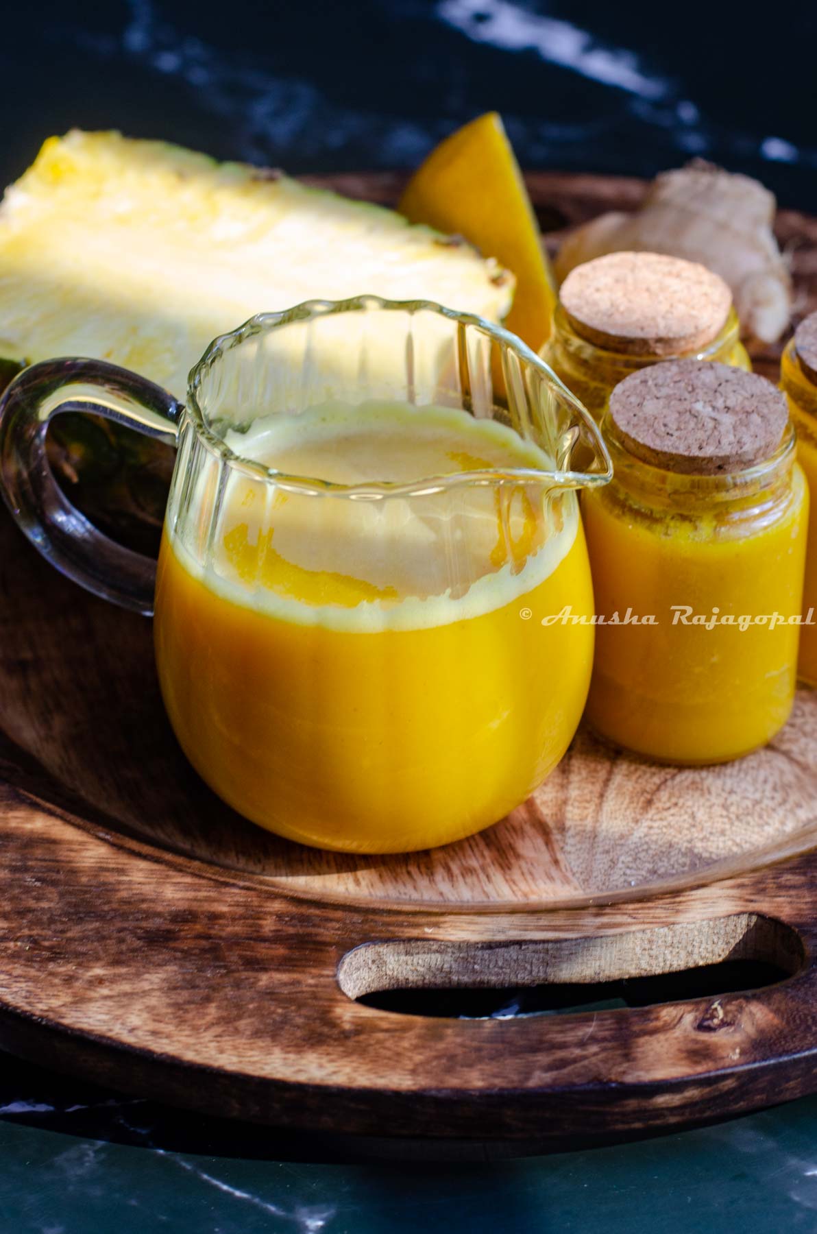 pineapple ginger shots in a small glass pitcher. Little jars of juice beside the tall glass. These little glasses have been sealed using a cork. A half cut pineapple and lemon at the back. All of these placed over a wooden plate.