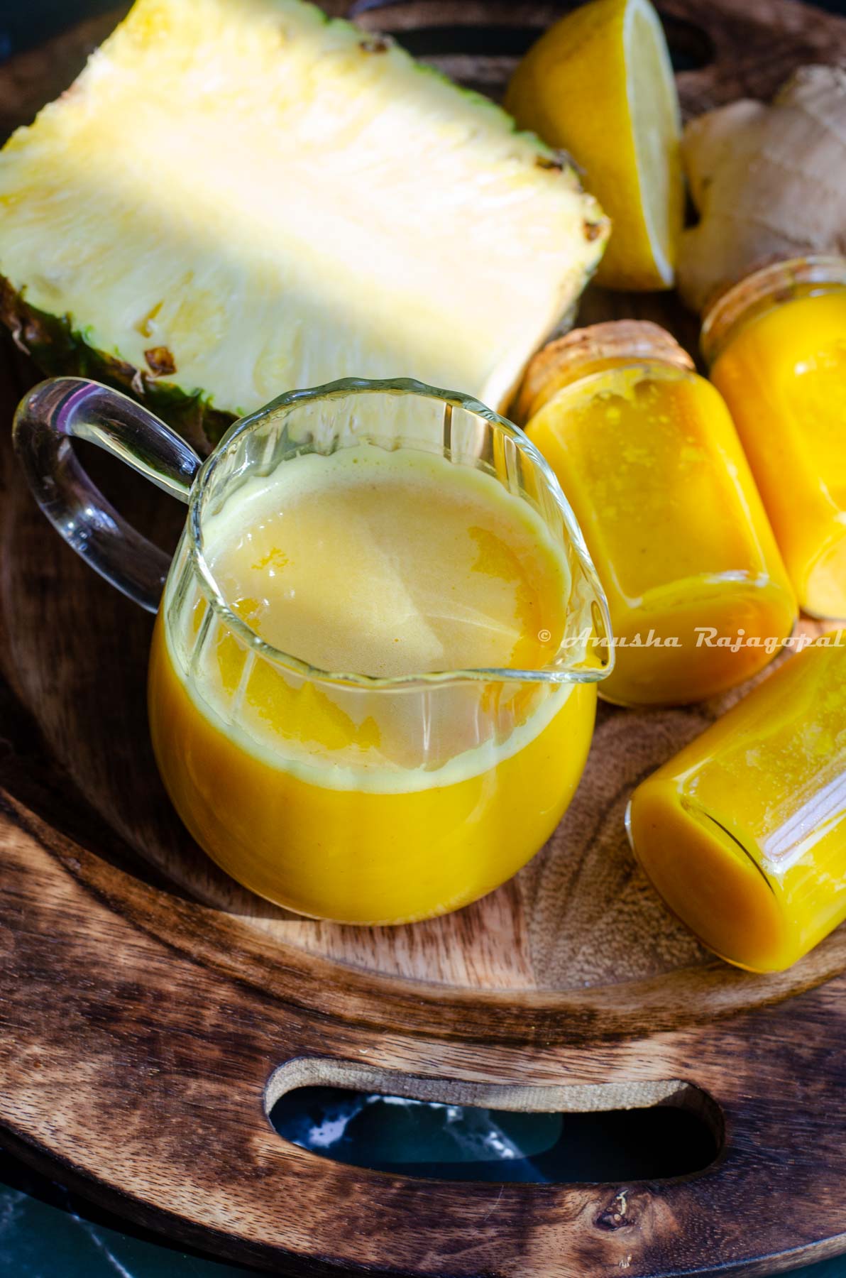 pineapple ginger shots in a small glass pitcher. Little jars of juice beside the tall glass. These little glasses have been sealed using a cork. A half cut pineapple and lemon at the back. All of these placed over a wooden plate.