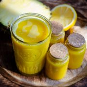 pineapple ginger juice served in a tall glass. Little jars of juice beside the tall glass. These little glasses have been sealed using a cork. A half cut pineapple and lemon at the back. All of these placed over a wooden plate.