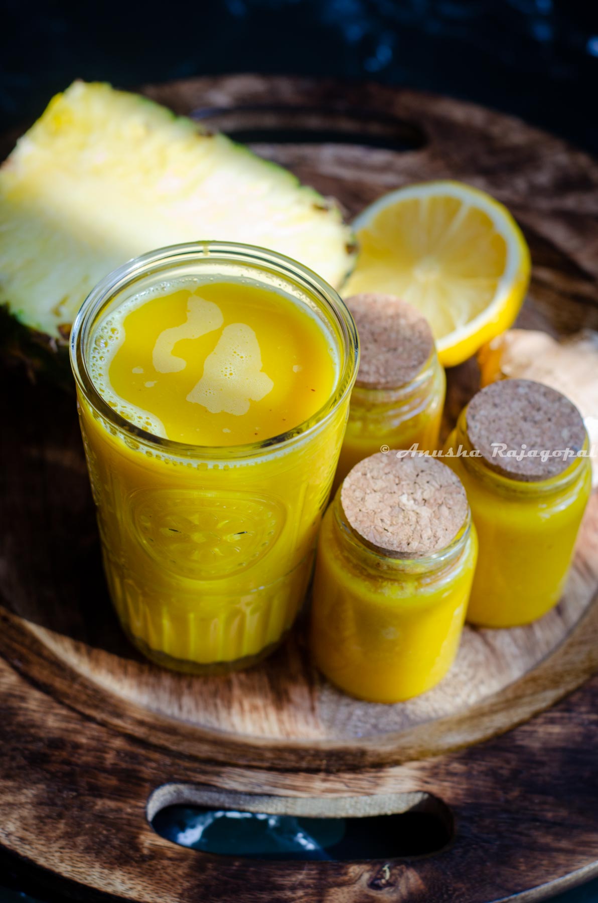 pineapple ginger juice served in a tall glass. Little jars of juice beside the tall glass. These little glasses have been sealed using a cork. A half cut pineapple and lemon at the back. All of these placed over a wooden plate.