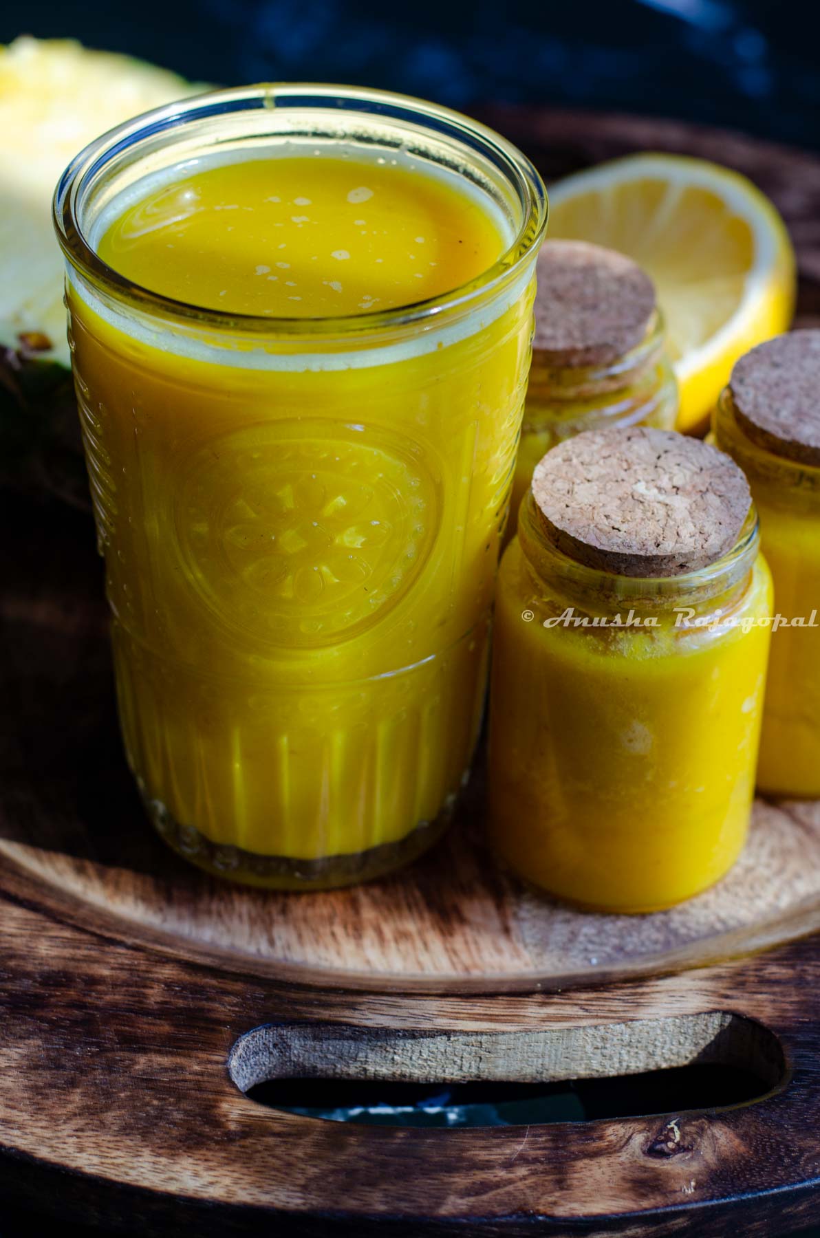 pineapple ginger juice served in a tall glass. Little jars of juice beside the tall glass. These little glasses have been sealed using a cork. A half cut pineapple and lemon at the back. All of these placed over a wooden plate.