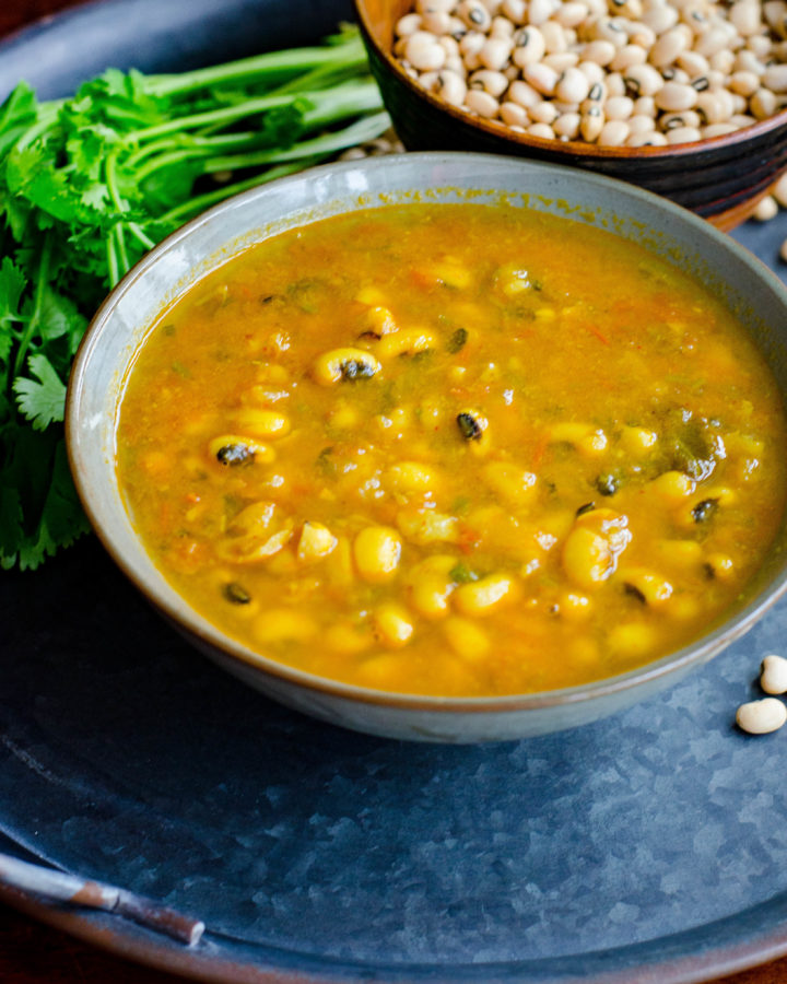 Black eyed peas curry served in a grey bowl placed on a blackish gray tray with scattered black eyed peas and cilantro at the background