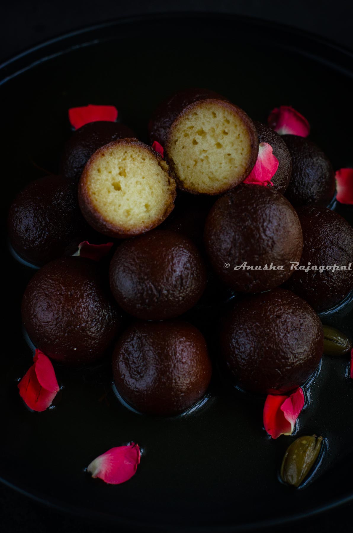 juicy kala jamuns and one cut up jamun stacked and served on a black plate. Garnished with rose petals.