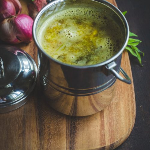 Onion rasam served in a stainless steel handled container placed on a wooden board