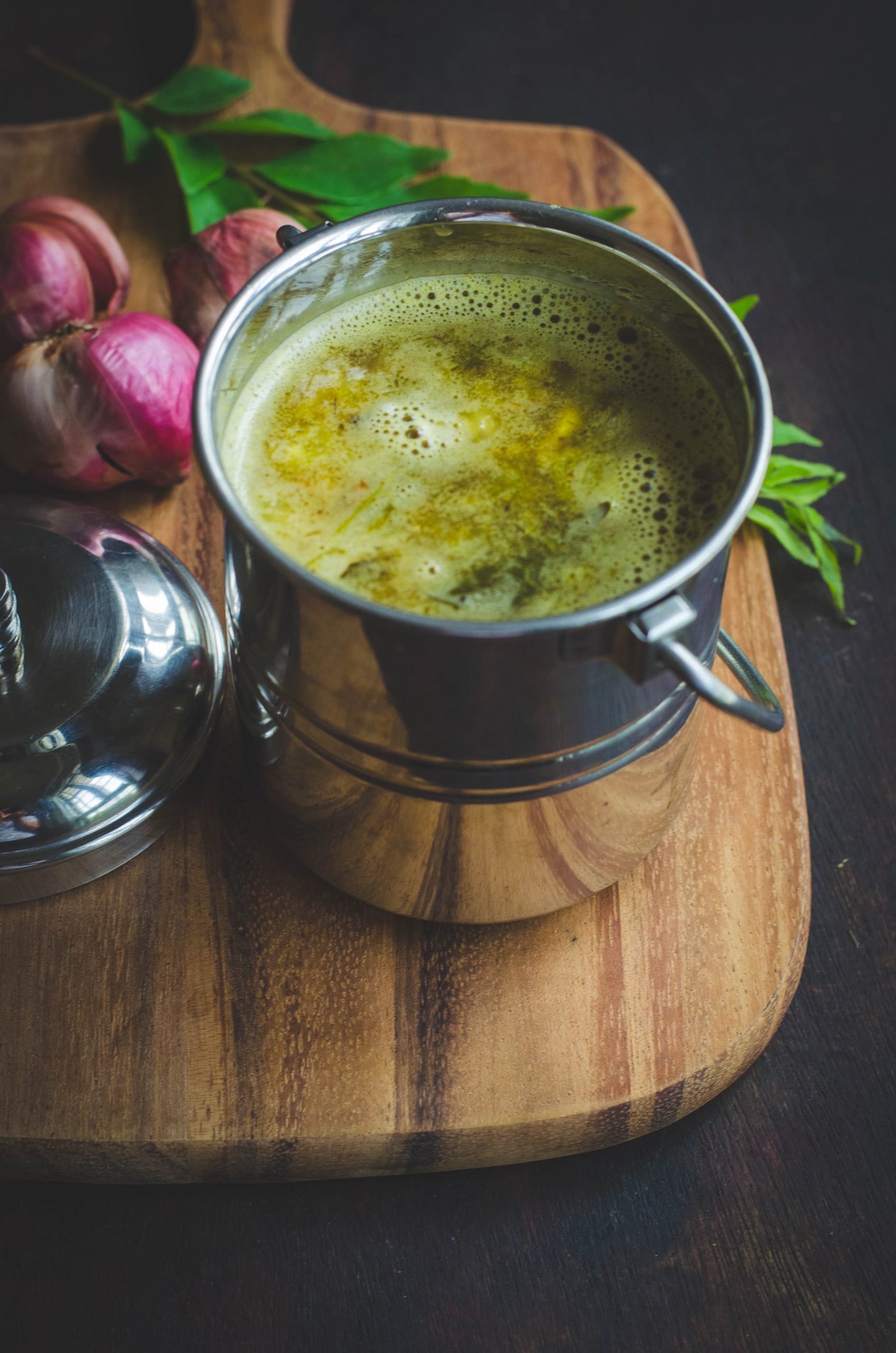 Onion rasam served in a stainless steel handled container placed on a wooden board