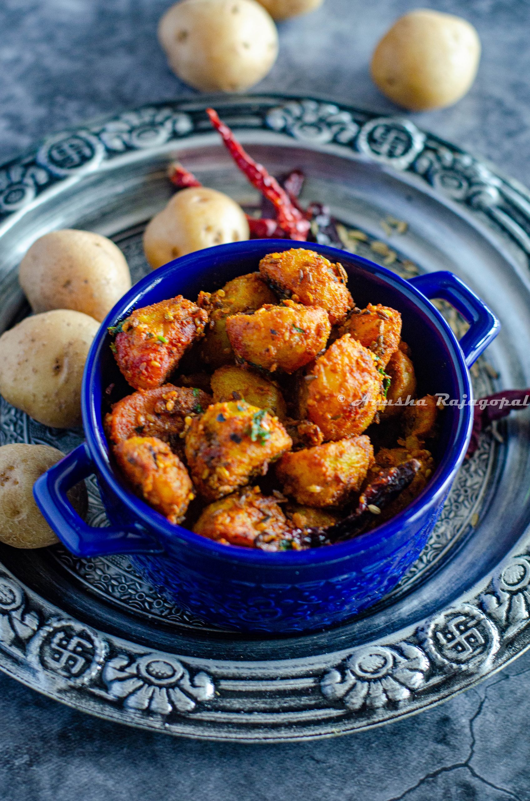 Jodhpuri aloo served in a blue casserole bowl placed over a silver plate, Baby potatoes at the background have been scattered