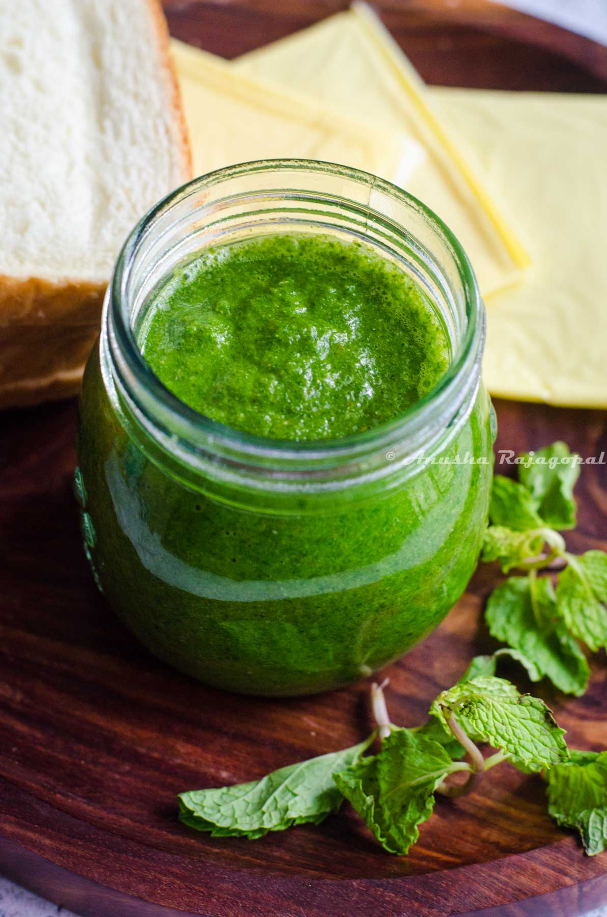 Bombay style sandwich chutney served in a glass jar with herbs, cheese and bread surrounding the serving platter.