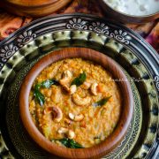 Instant Pot Bisi Bele Bath served in a wooden bowl with garnishing of curry leaves and cashews. The bowl is placed over an antique platter and is surrounded by bowls of raita and fried okra.