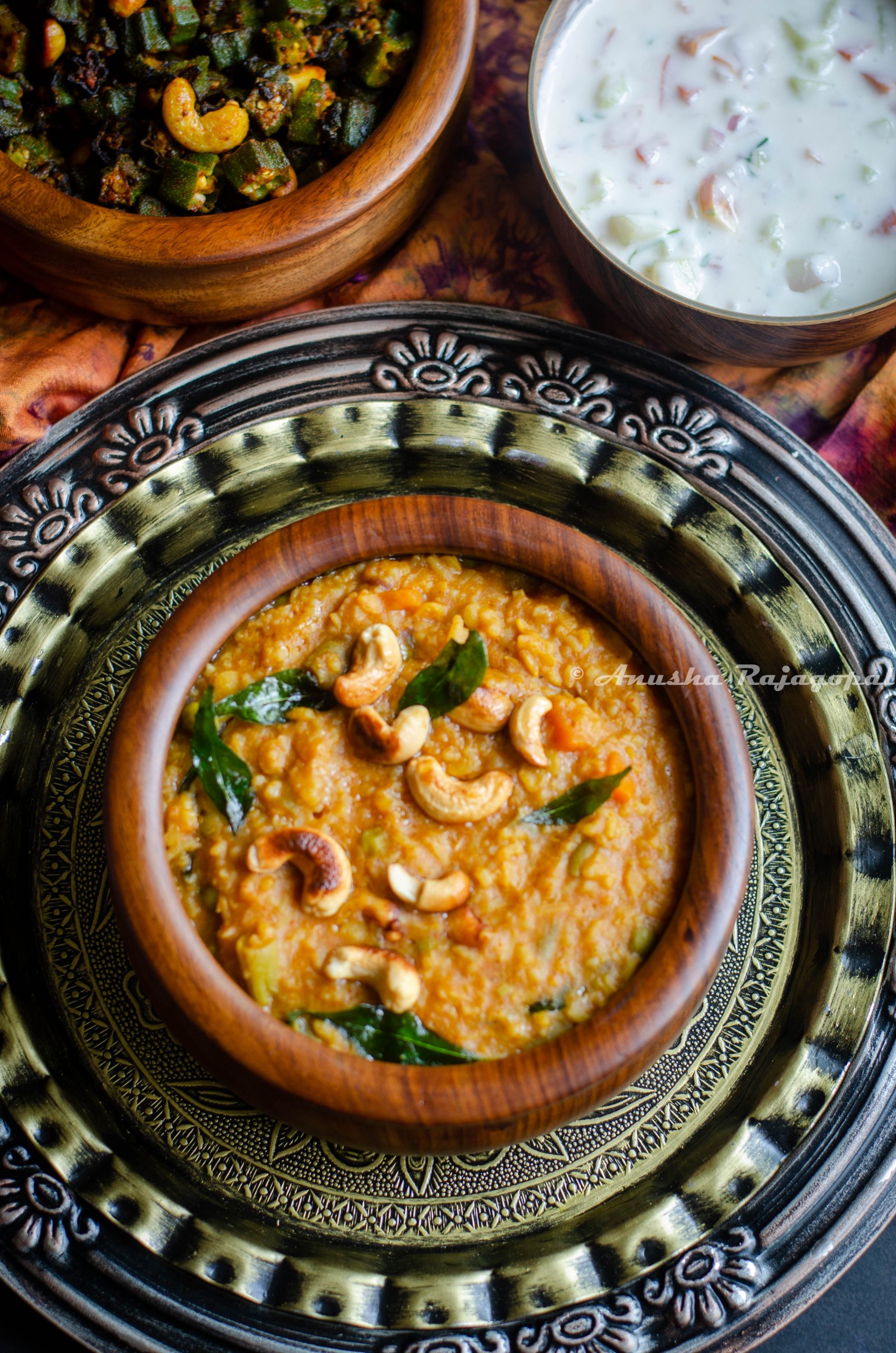 Instant Pot Bisi Bele Bath served in a wooden bowl with garnishing of curry leaves and cashews. The bowl is placed over an antique platter and is surrounded by bowls of raita and fried okra.