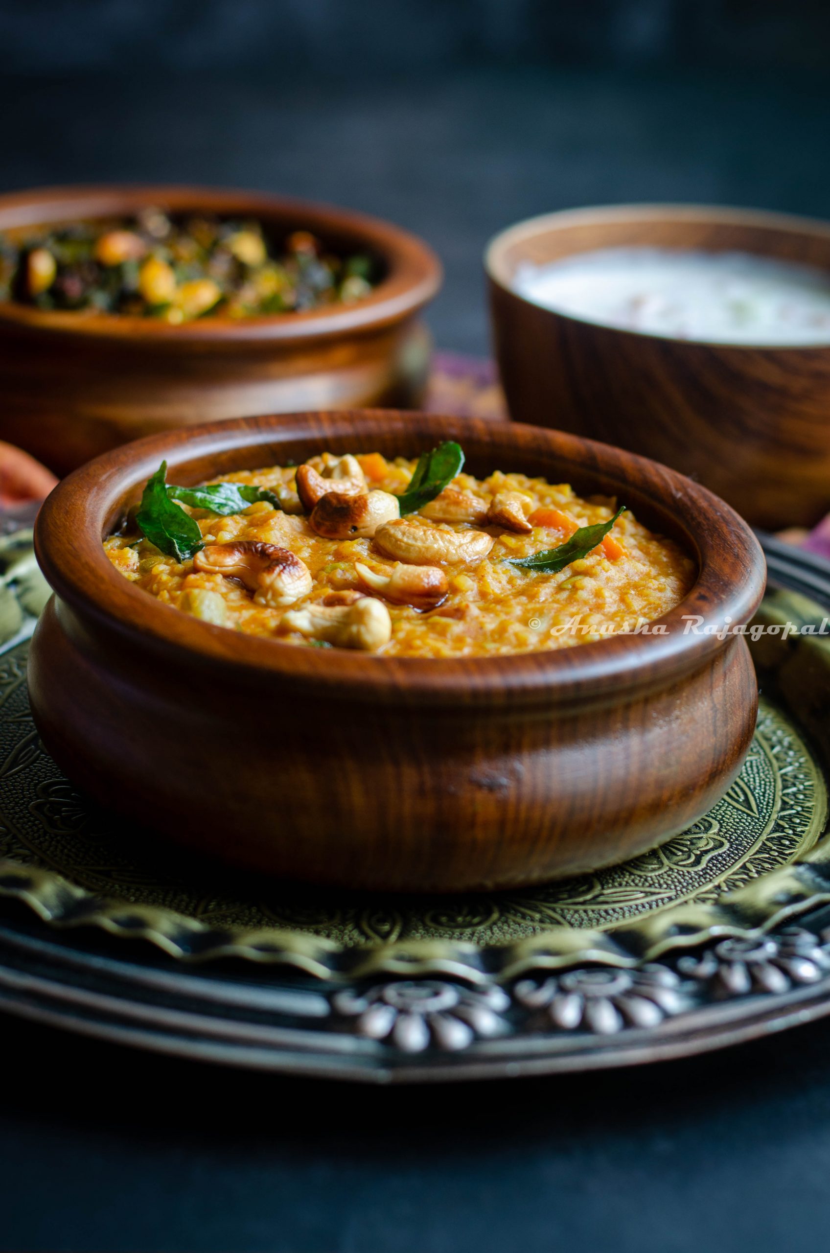 Instant Pot Bisi Bele Bath served in a wooden bowl with garnishing of curry leaves and cashews. The bowl is placed over an antique platter and is surrounded by bowls of raita and fried okra.