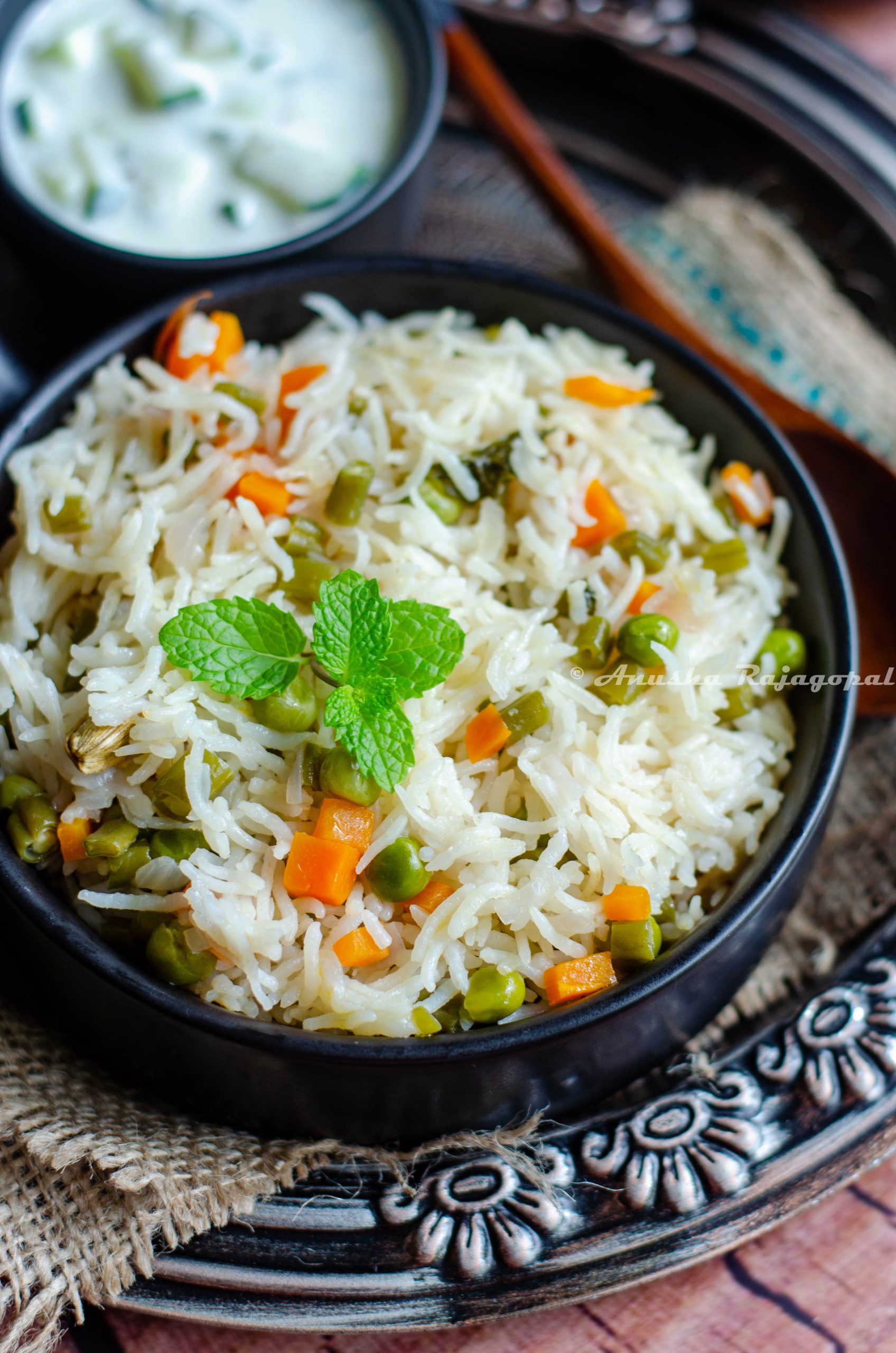 instant pot vegetable pulao in a black bowl served with cucumber raita. both bowls placed on a burlap mat over a rustic platter.