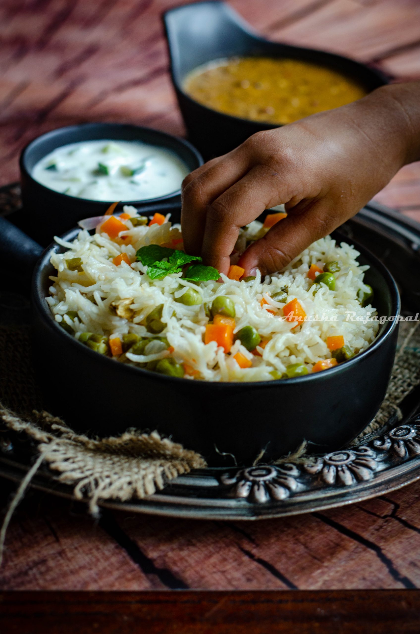 instant pot vegetable pulao in a black bowl served with cucumber raita. both bowls placed on a burlap mat over a rustic platter. A kid reaches out to take some carrots off the bowl.