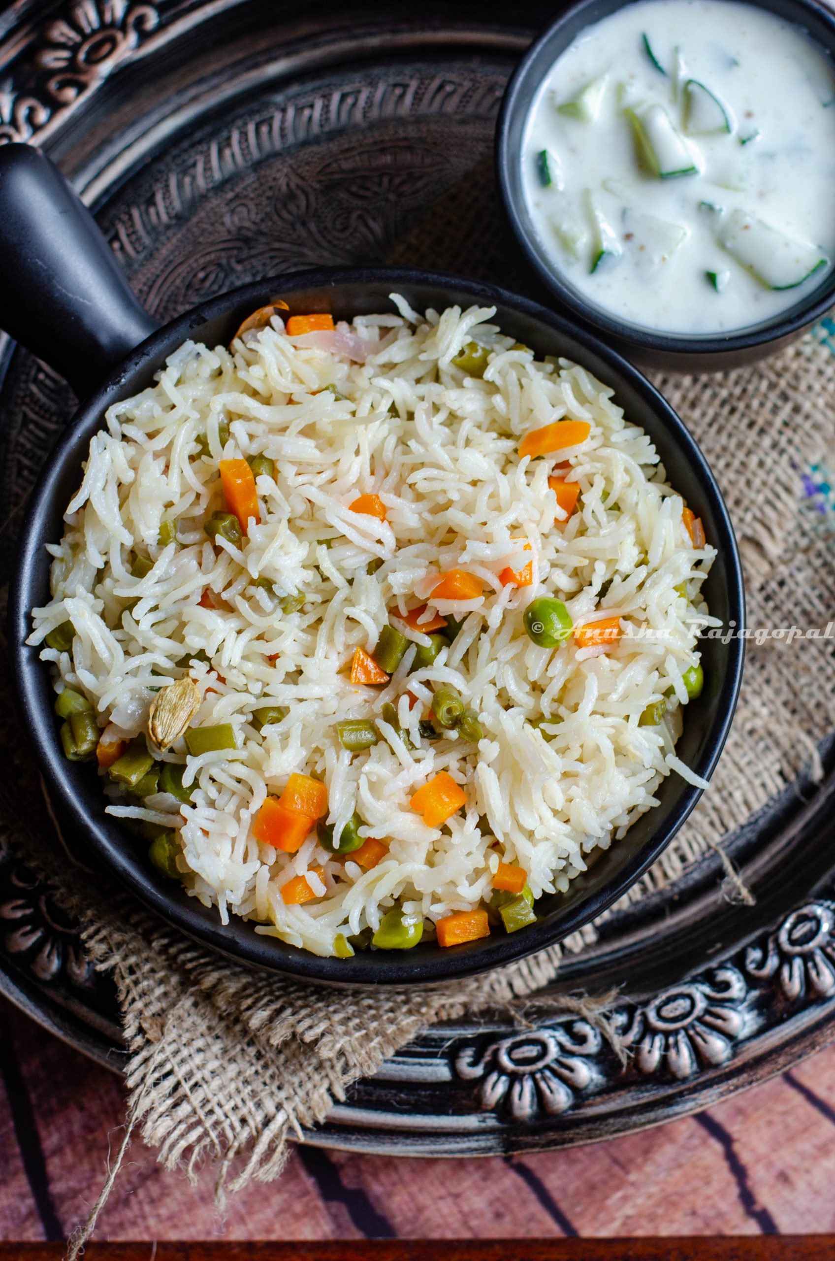 instant pot vegetable pulao in a black bowl served with cucumber raita. both bowls placed on a burlap mat over a rustic platter.