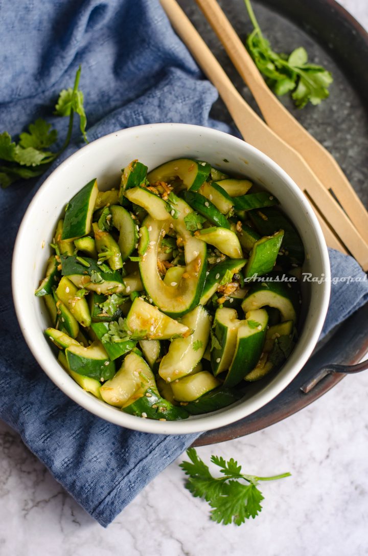 chinese cucumber salad served in a white bowl placed on a blue napkin. Cilantro and salad spoons by the side.