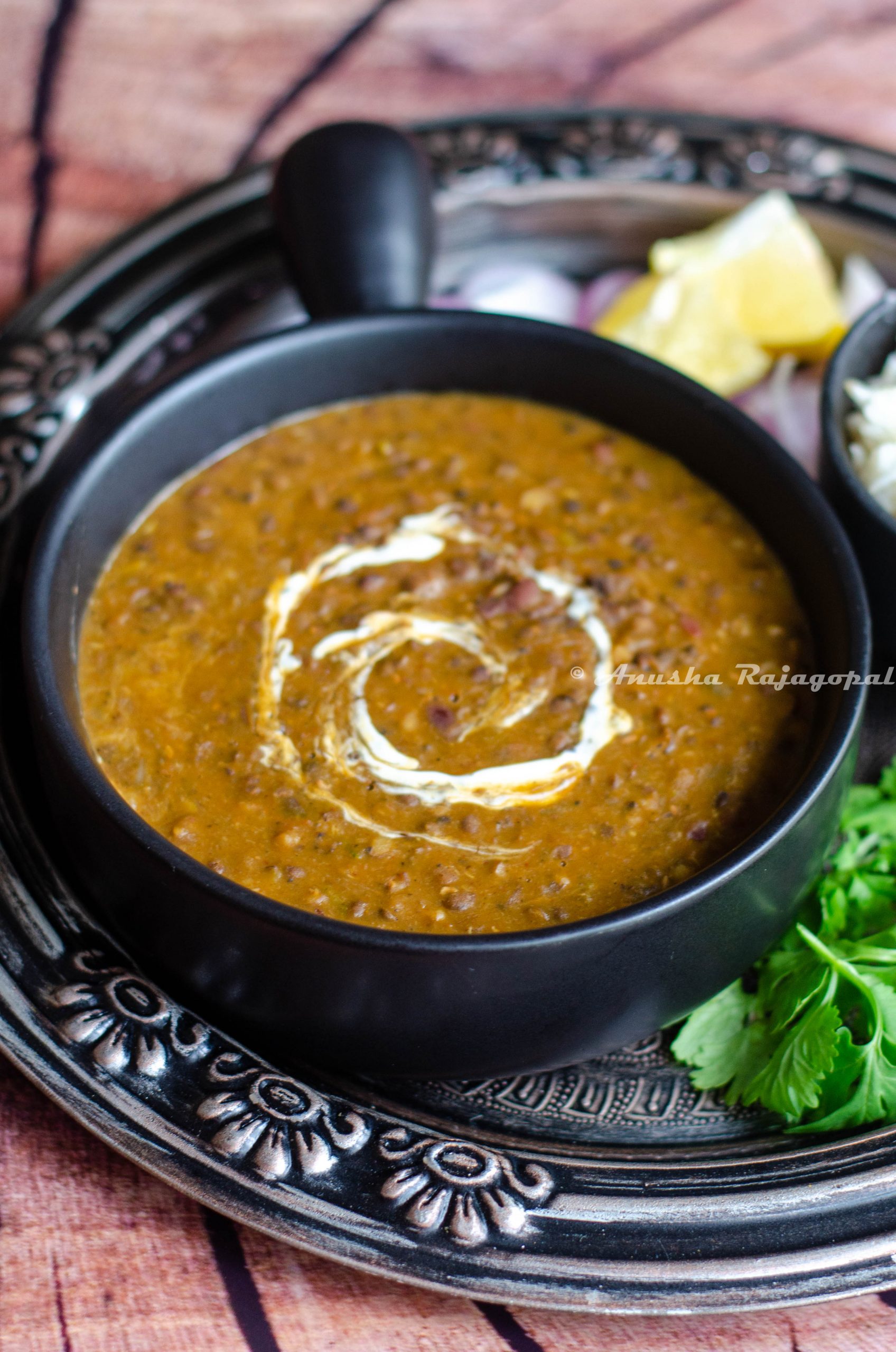 instant pot vegan dal makhani served in a black ladled bowl placed on an antique plate. Lemon wedges and onions with cumin rice by the side.