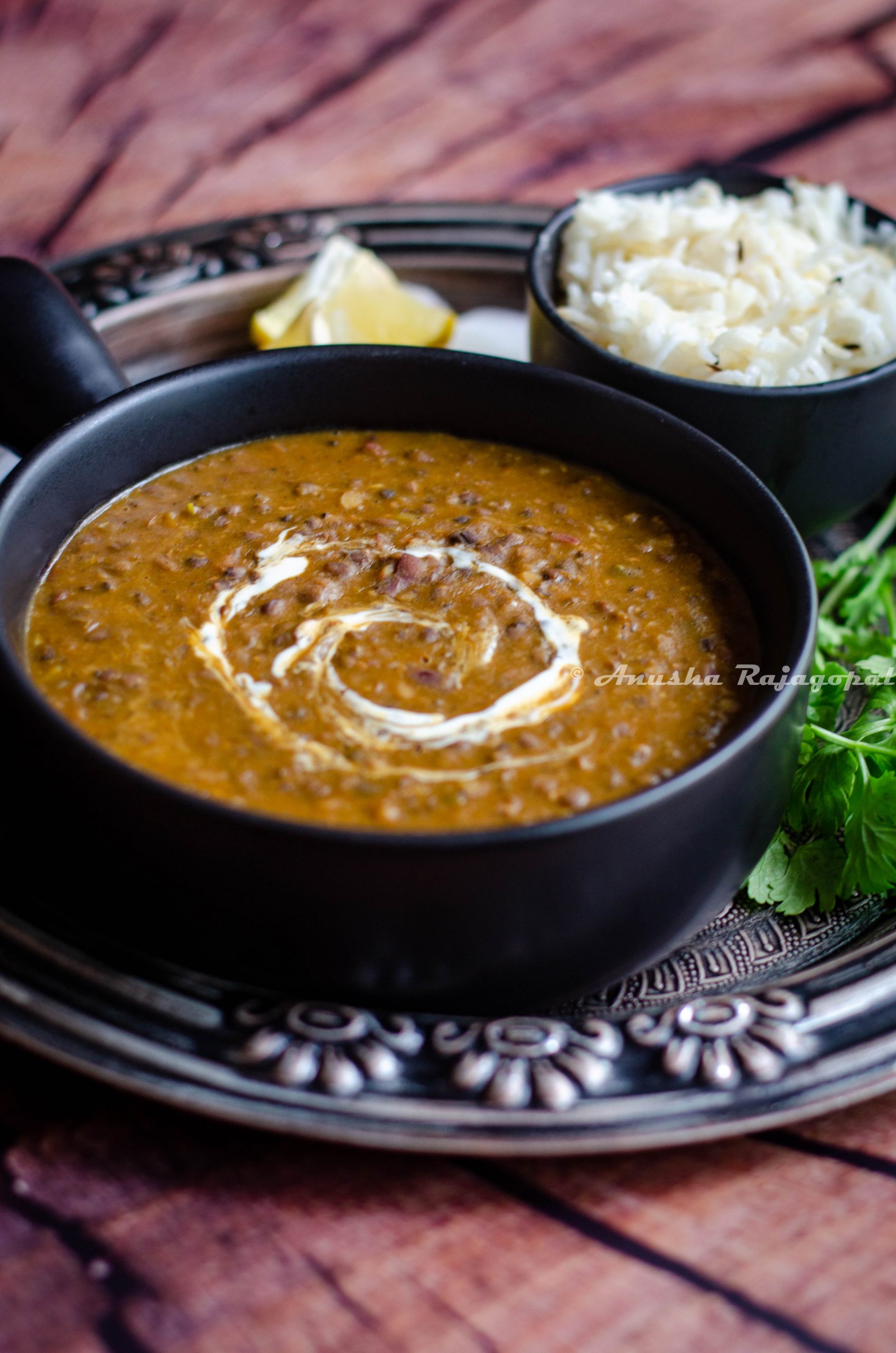 instant pot vegan dal makhani served in a black ladled bowl placed on an antique plate. Lemon wedges and onions with cumin rice by the side.