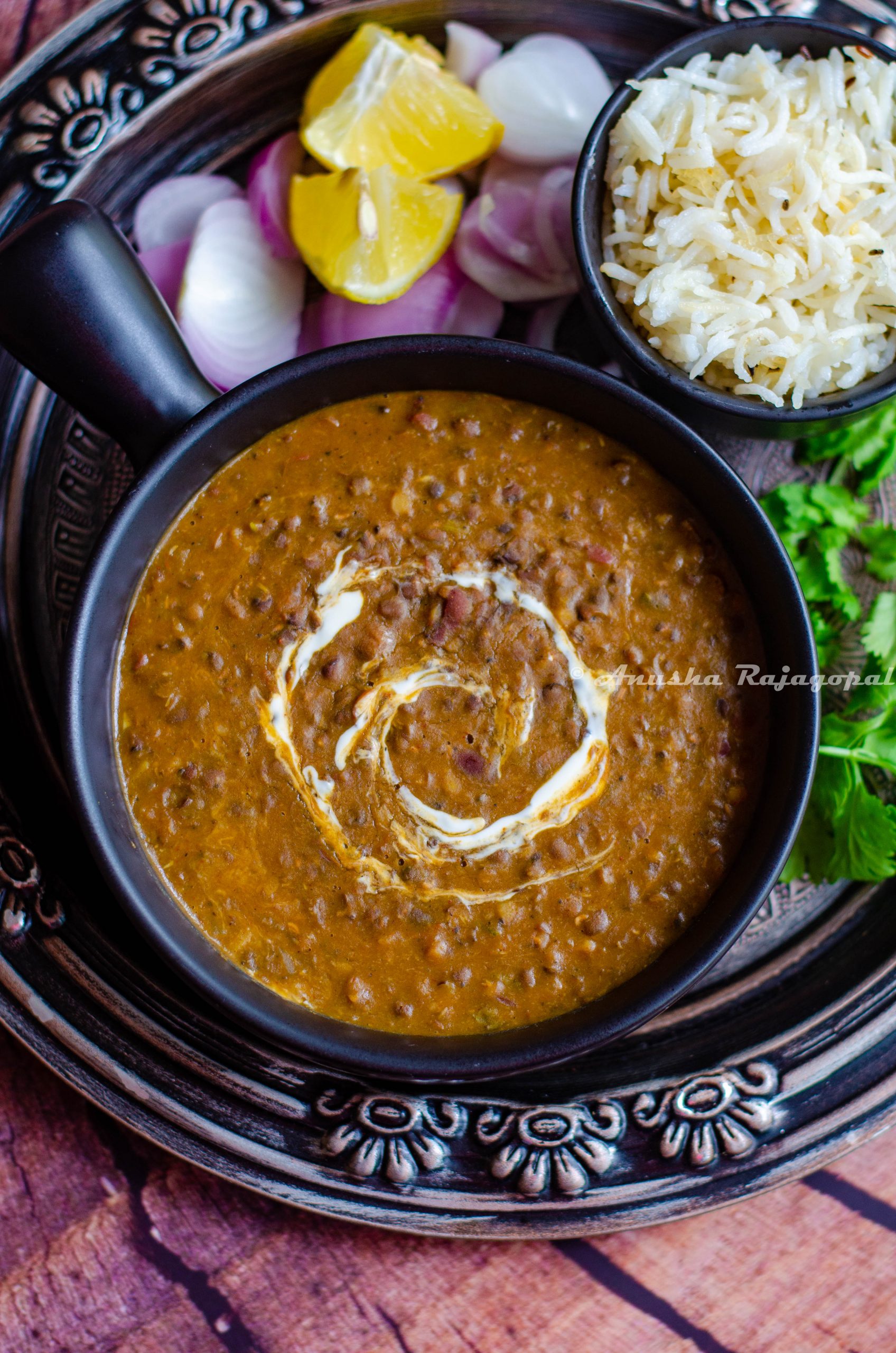 instant pot vegan dal makhani served in a black ladled bowl placed on an antique plate. Lemon wedges and onions with cumin rice by the side.