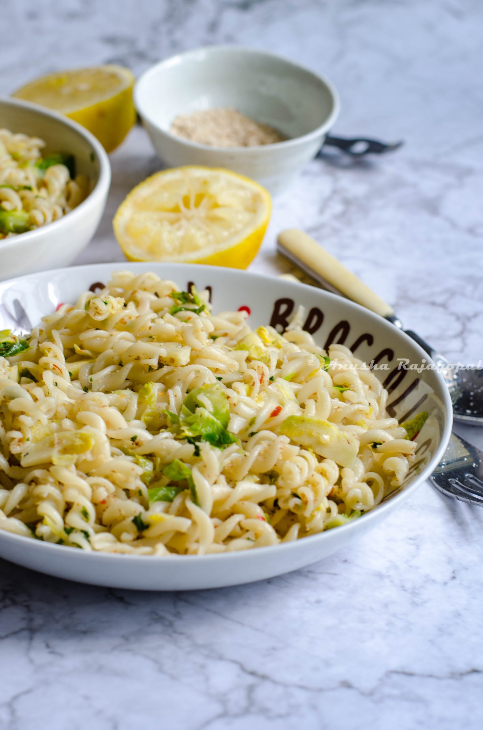 vegan lemon brussel sprouts pasta served in a white plate set on a white marble background. Forks, spoons, lemon and ground almonds by the side.