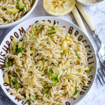 vegan lemon brussel sprouts pasta served in a white plate set on a white marble background. Forks, spoons, lemon and ground almonds by the side.