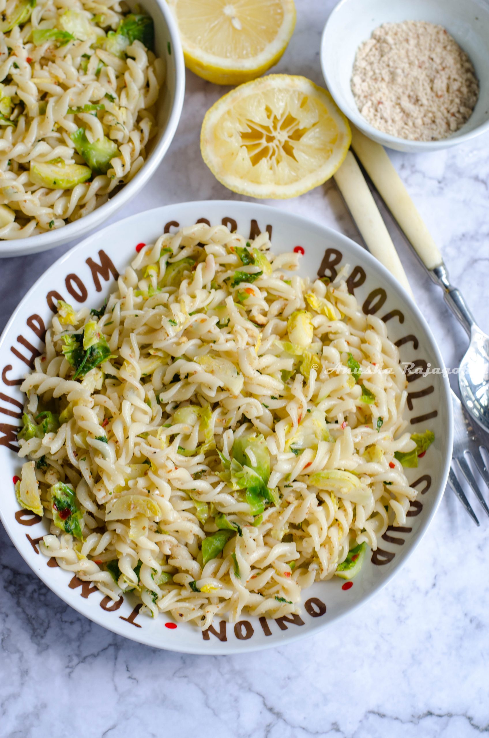 vegan lemon brussel sprouts pasta served in a white plate set on a white marble background. Forks, spoons, lemon and ground almonds by the side.