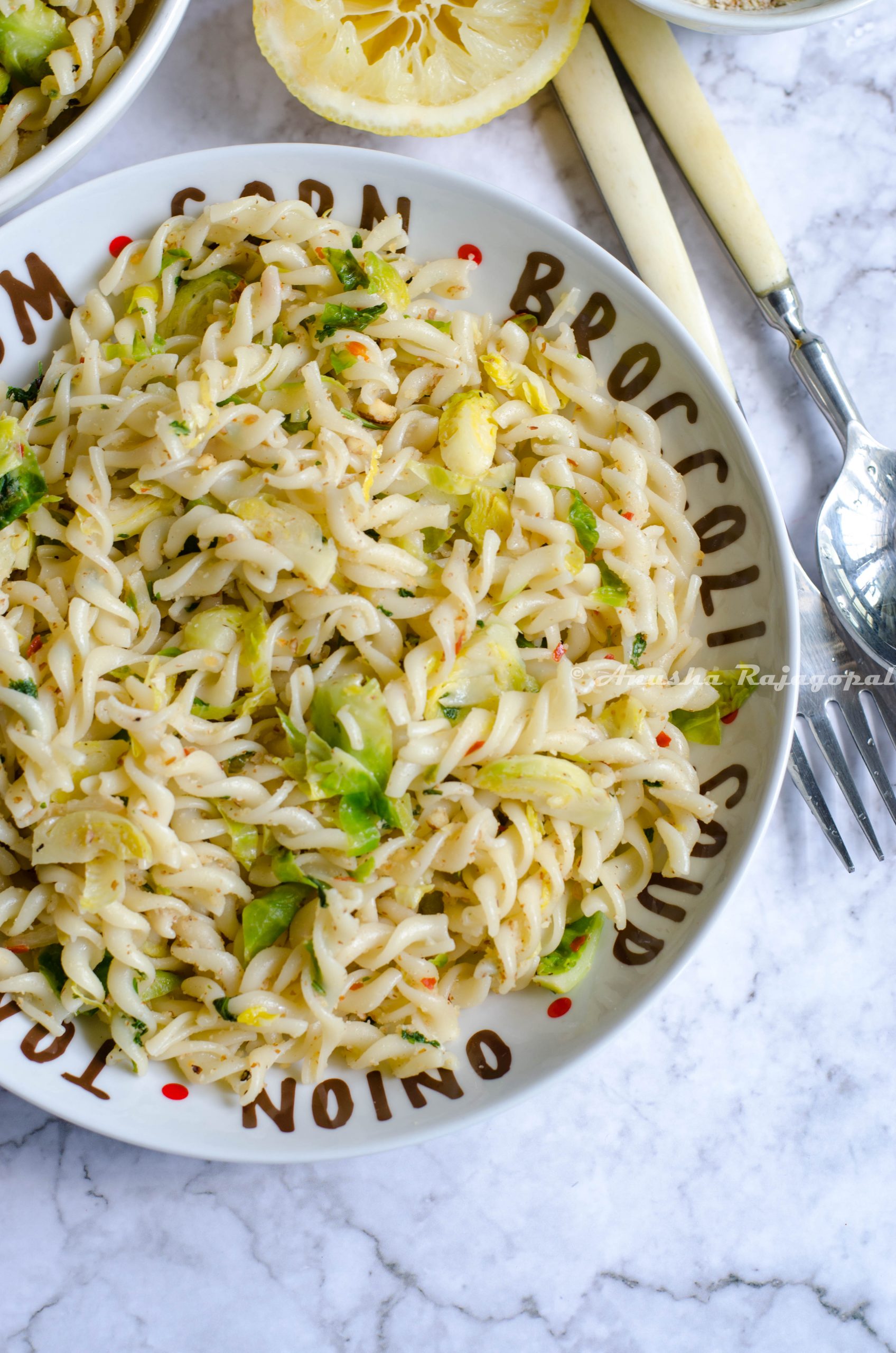 vegan lemon brussel sprouts pasta served in a white plate set on a white marble background. Forks, spoons, lemon and ground almonds by the side.