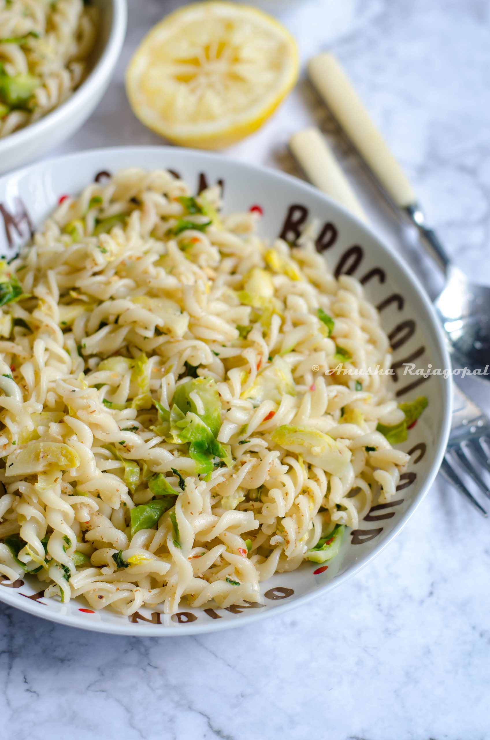 vegan lemon brussel sprouts pasta served in a white plate set on a white marble background. Forks, spoons, lemon and ground almonds by the side.