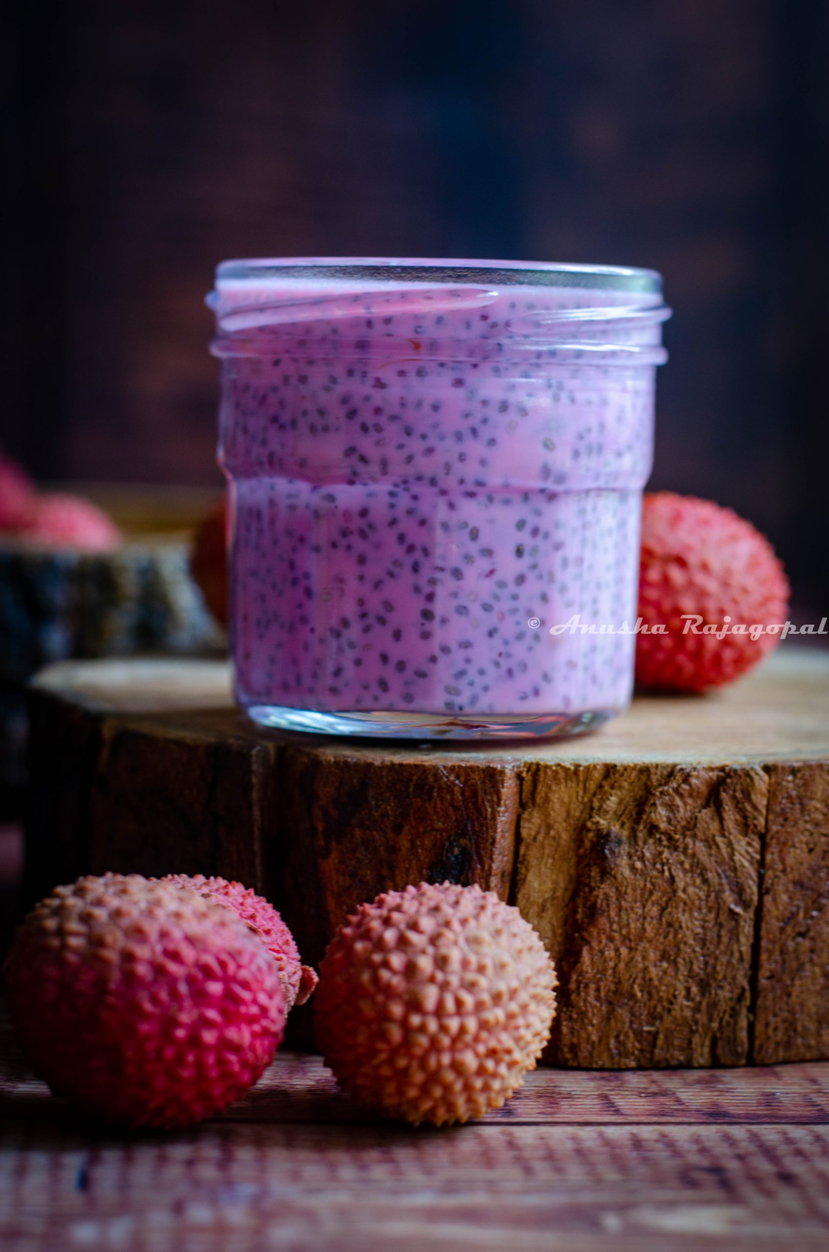 lychee rose chia pudding in a glass jar perched on a wooden tray, lychees scattered around