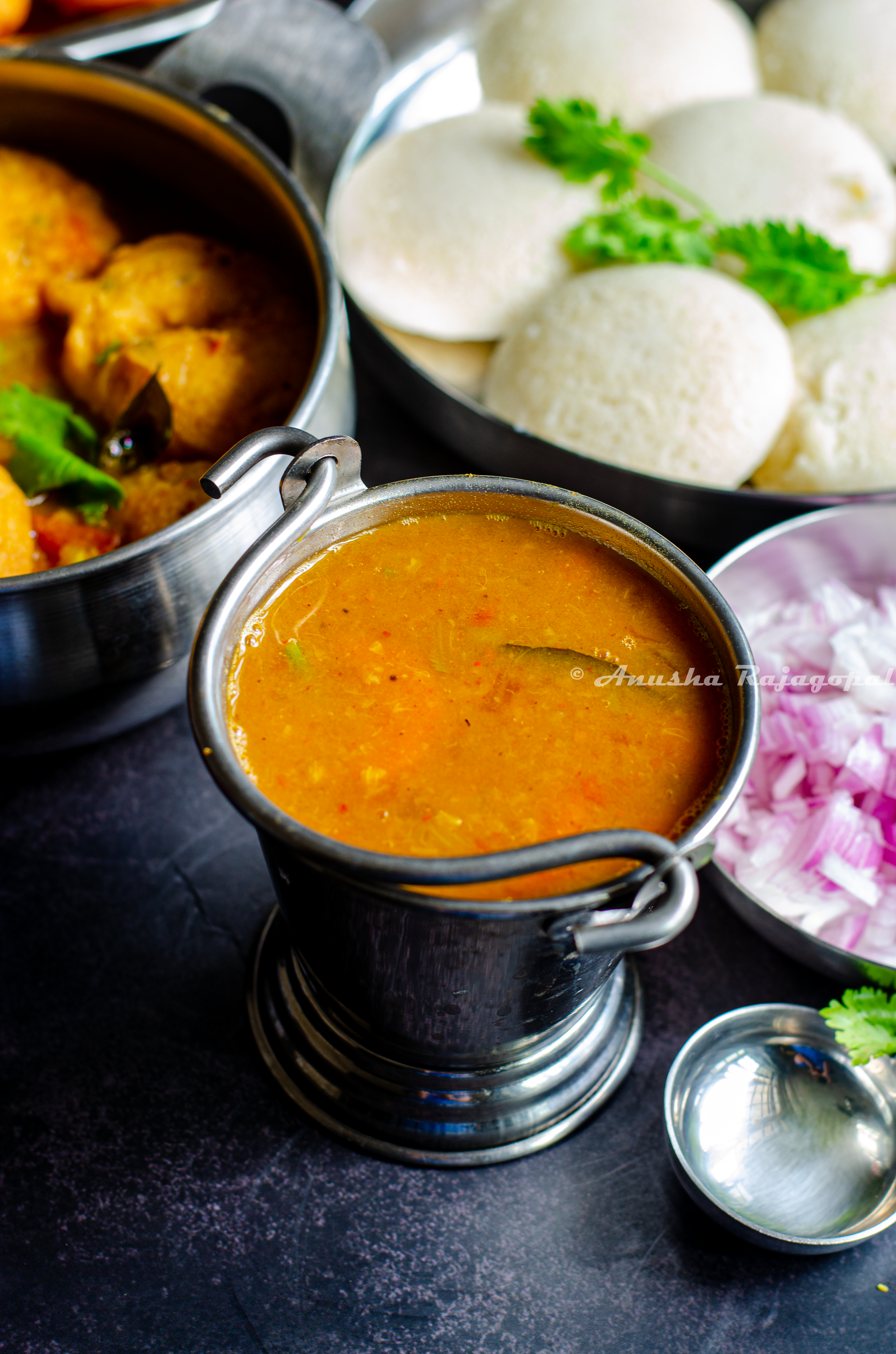 hotel style tiffin sambar served in a pail with idli, vada,chopped onions and a ladle by the side.