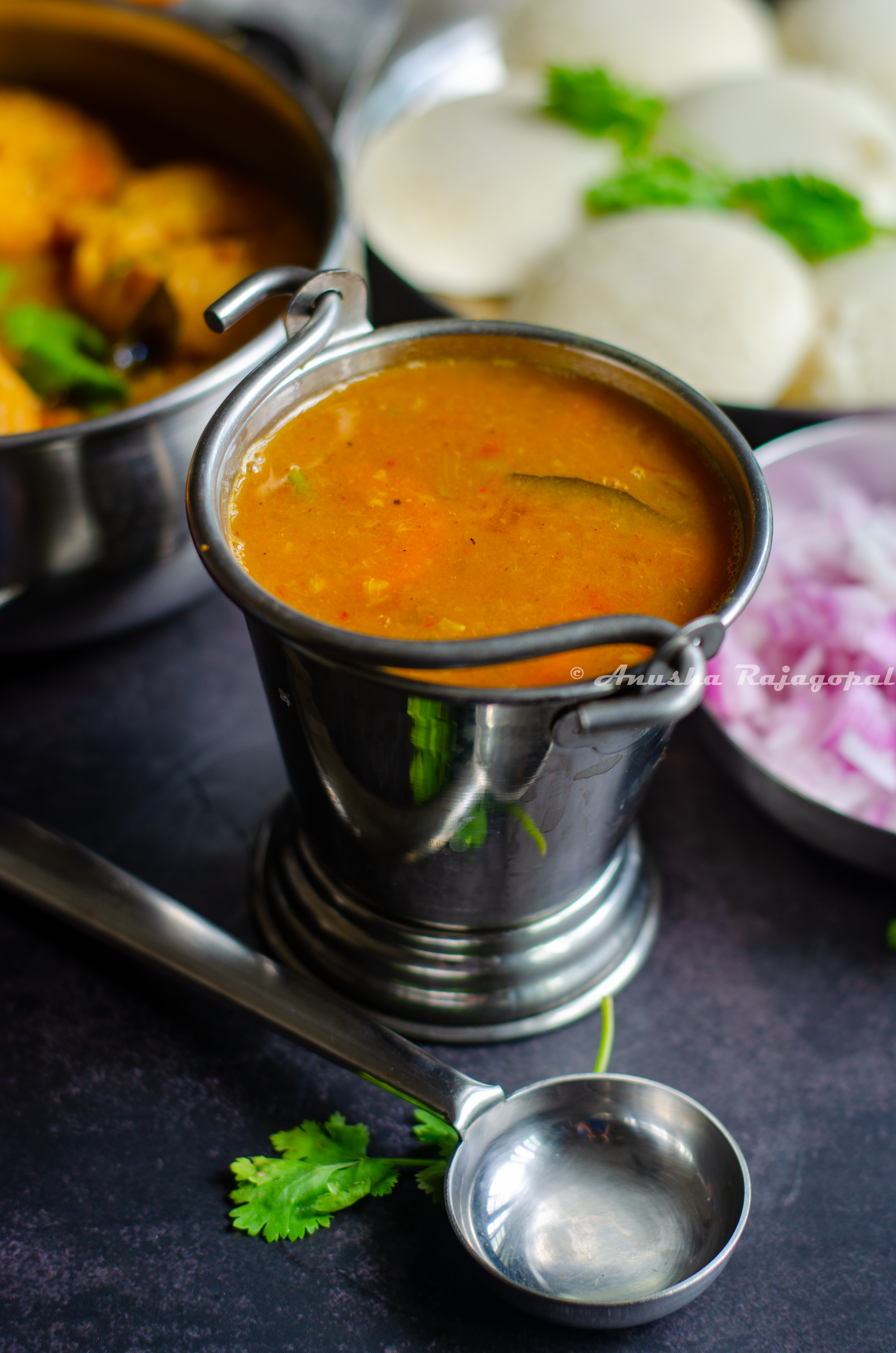 hotel style tiffin sambar served in a pail with idli, vada,chopped onions and a ladle by the side.