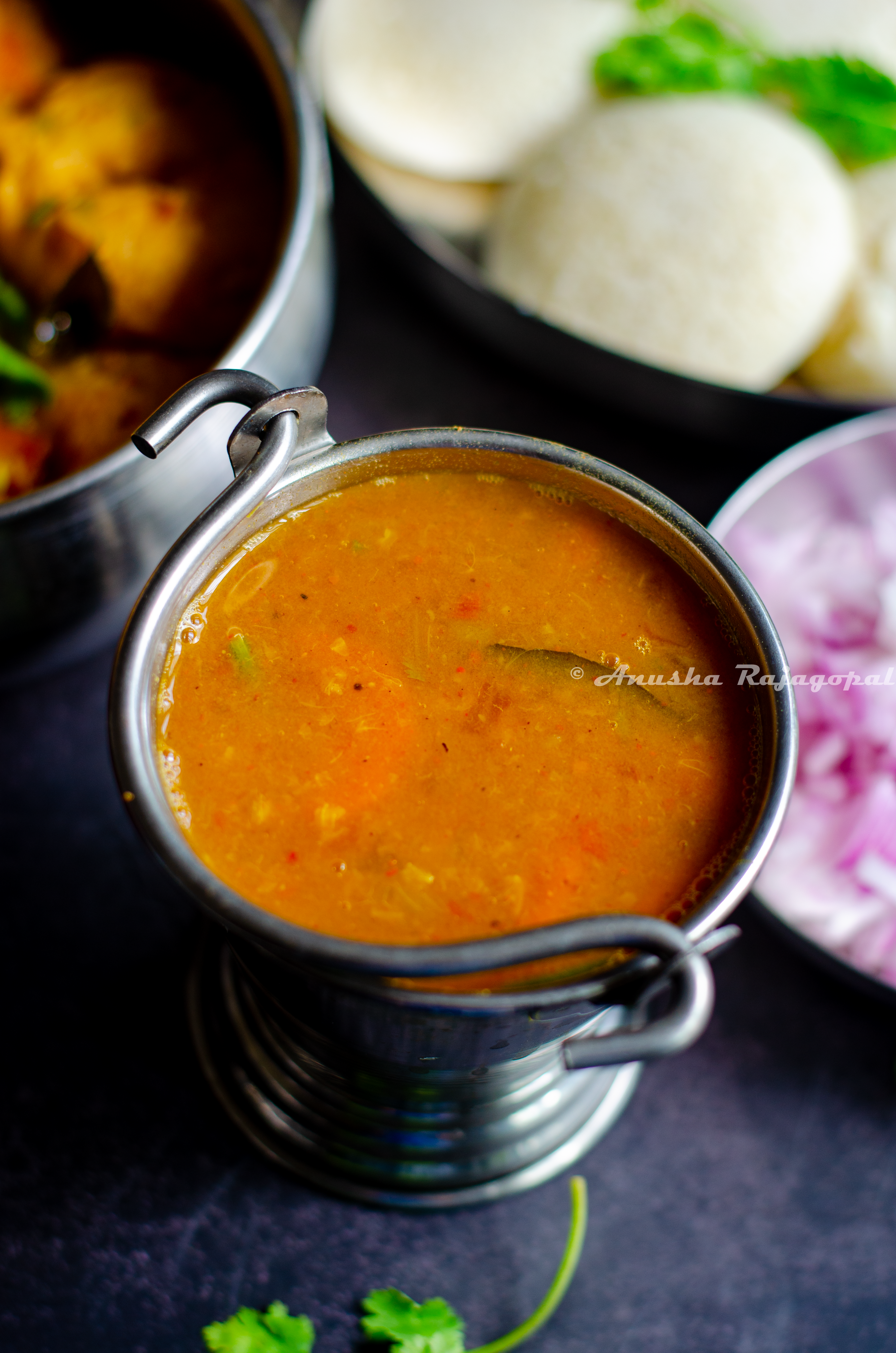hotel style tiffin sambar served in a pail with idli, vada,chopped onions and a ladle by the side.