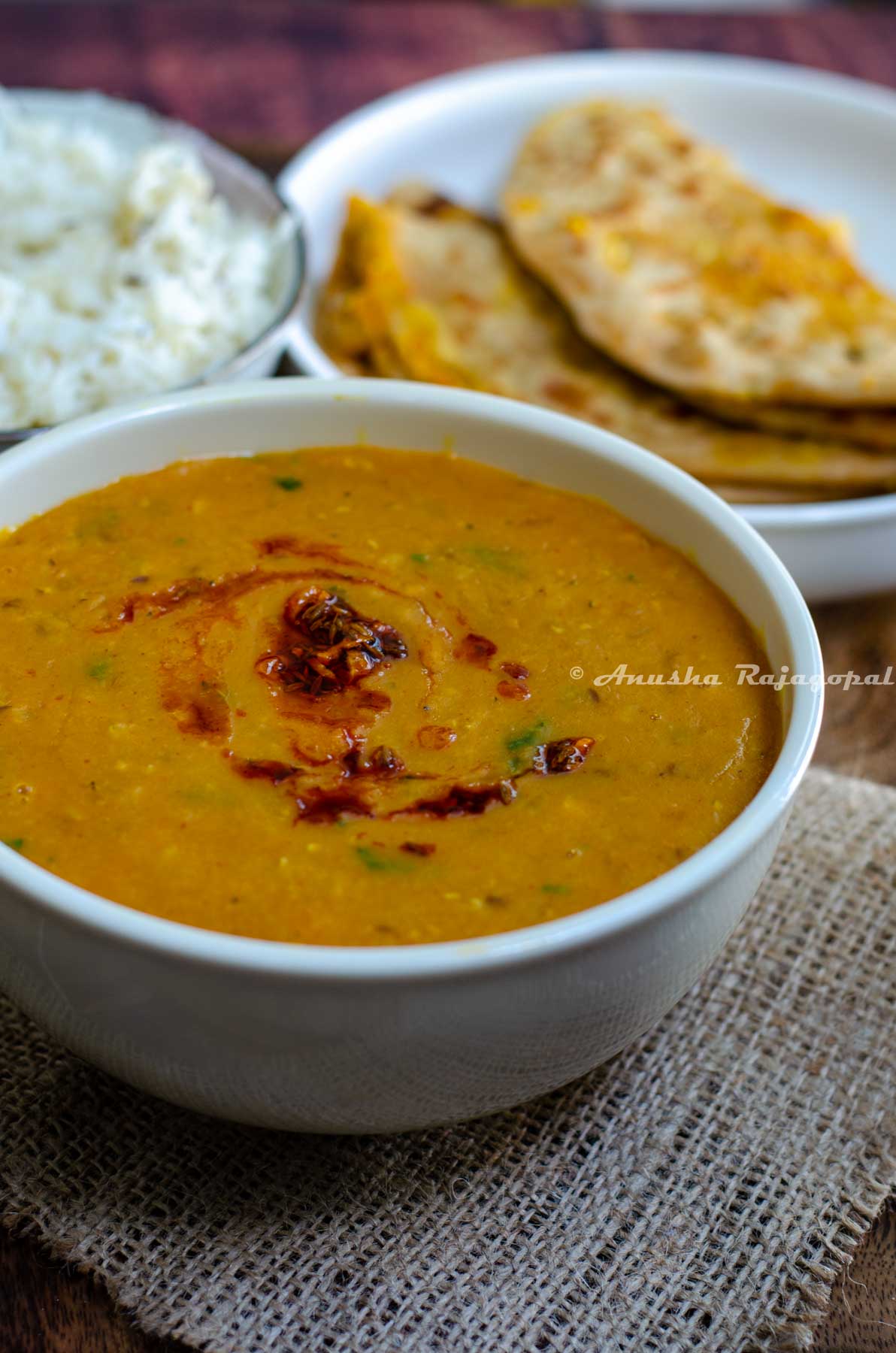 instant pot vegan dahl served in a white bowl placed on a burlap mat. Dahl topped with a garlic and paprika tempering. Jeera rice and Gobi paratha at the background.