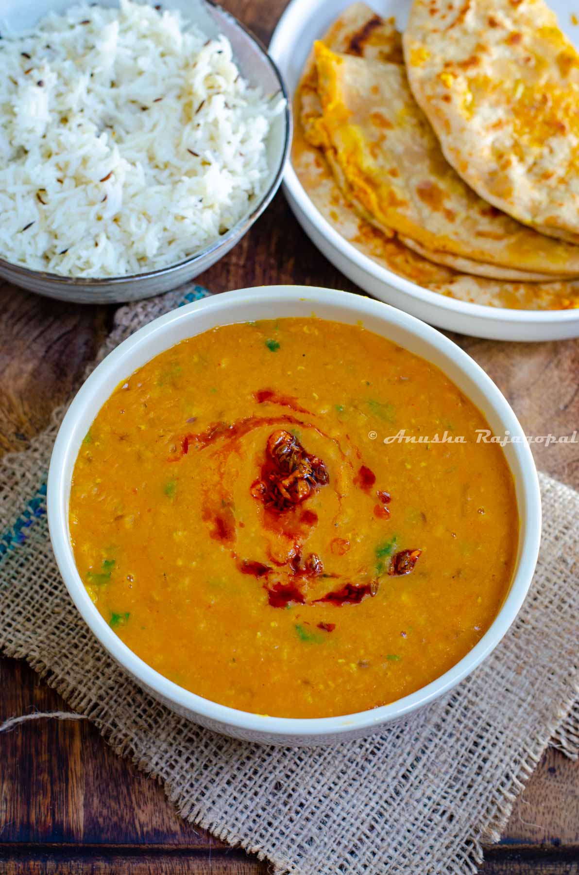 instant pot vegan dahl served in a white bowl placed on a burlap mat. Dahl topped with a garlic and paprika tempering. Jeera rice and Gobi paratha at the background.