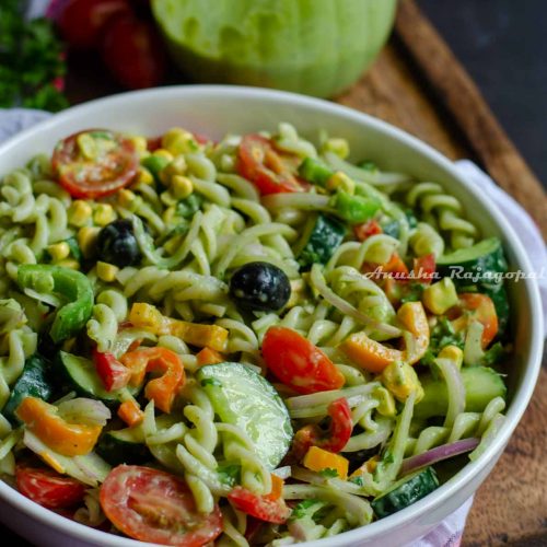 summer pasta salad served in a deep plate placed on a white tea towel. Dressing jar at the back and some tomatoes and herbs by the side.