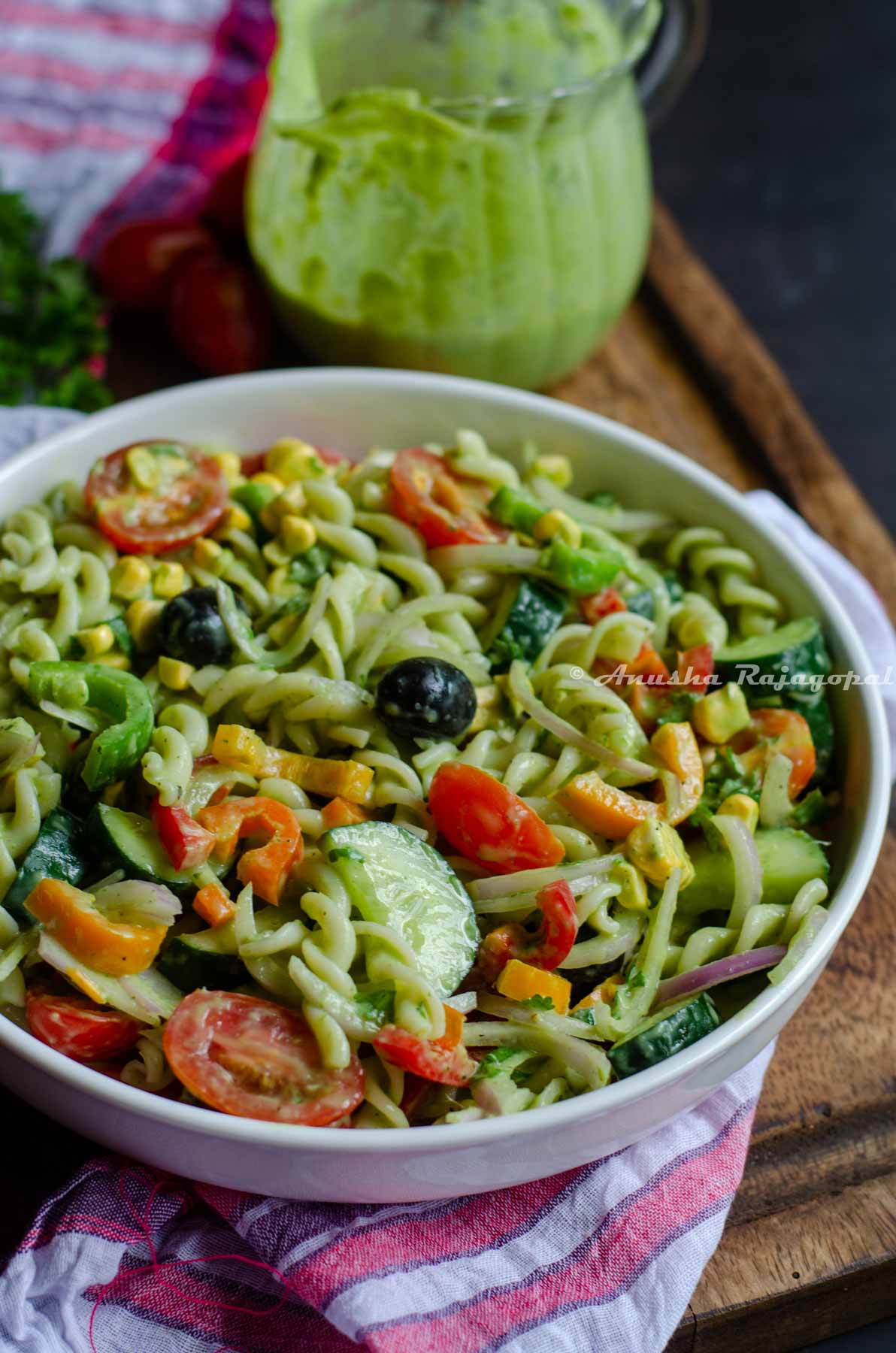 summer pasta salad served in a deep plate placed on a white tea towel. Dressing jar at the back and some tomatoes and herbs by the side.